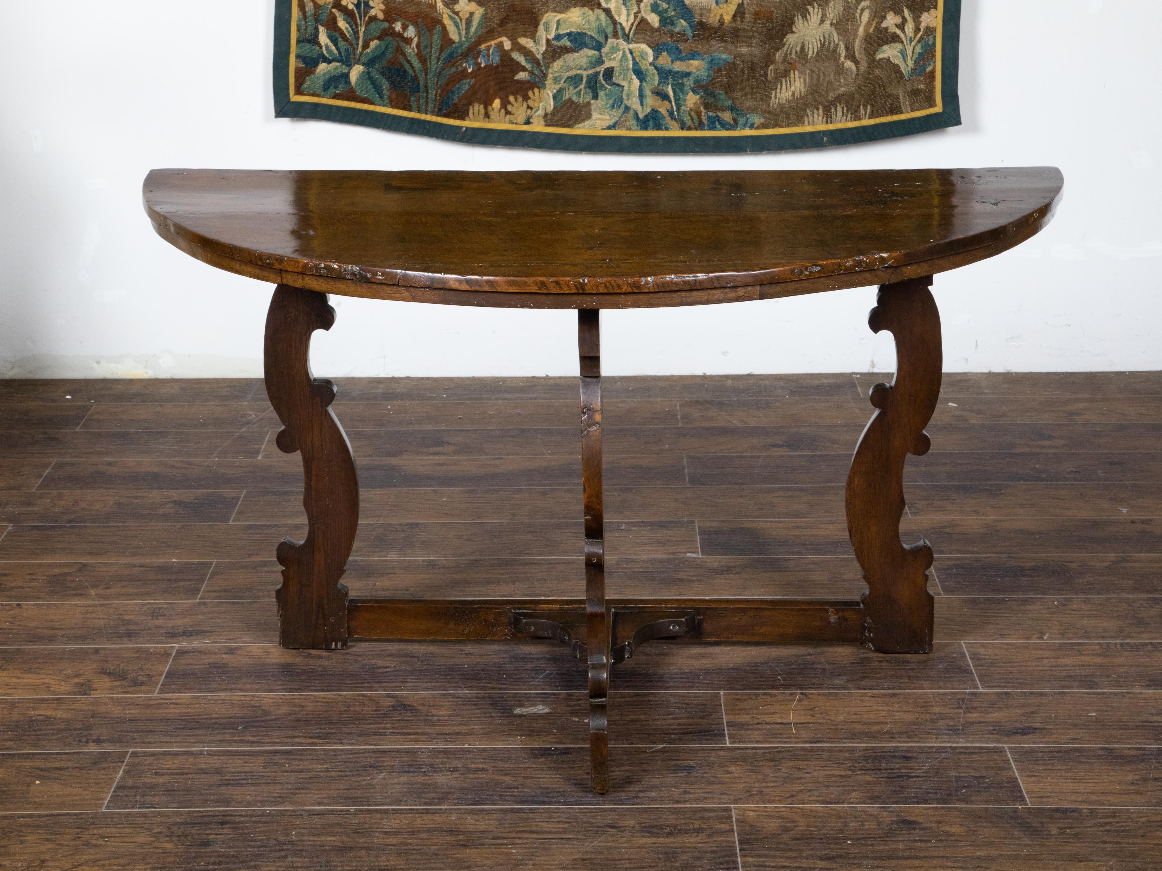 A large Italian Baroque style dark walnut demi lune console table from the 18th century, with semi-circular top, carved legs and cross stretcher. Created in Italy during the 18th century, this walnut demi lune table features a semi-circular planked