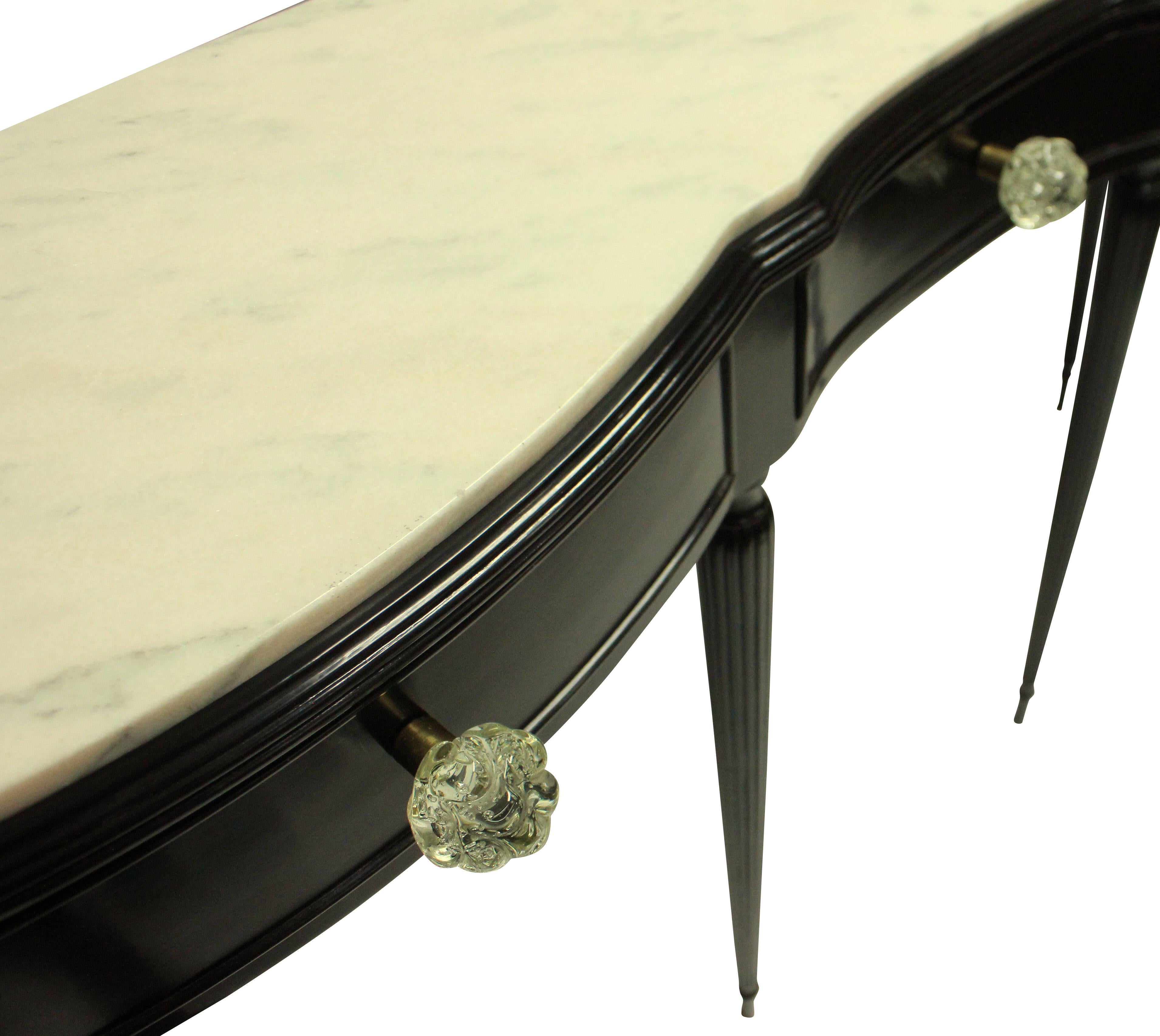 A large Italian midcentury ebonized console table with a serpentine shape upon tapering fluted legs. The three drawers have beautiful hand blown handles, full of air bubbles and the top has a cream inset marble top.