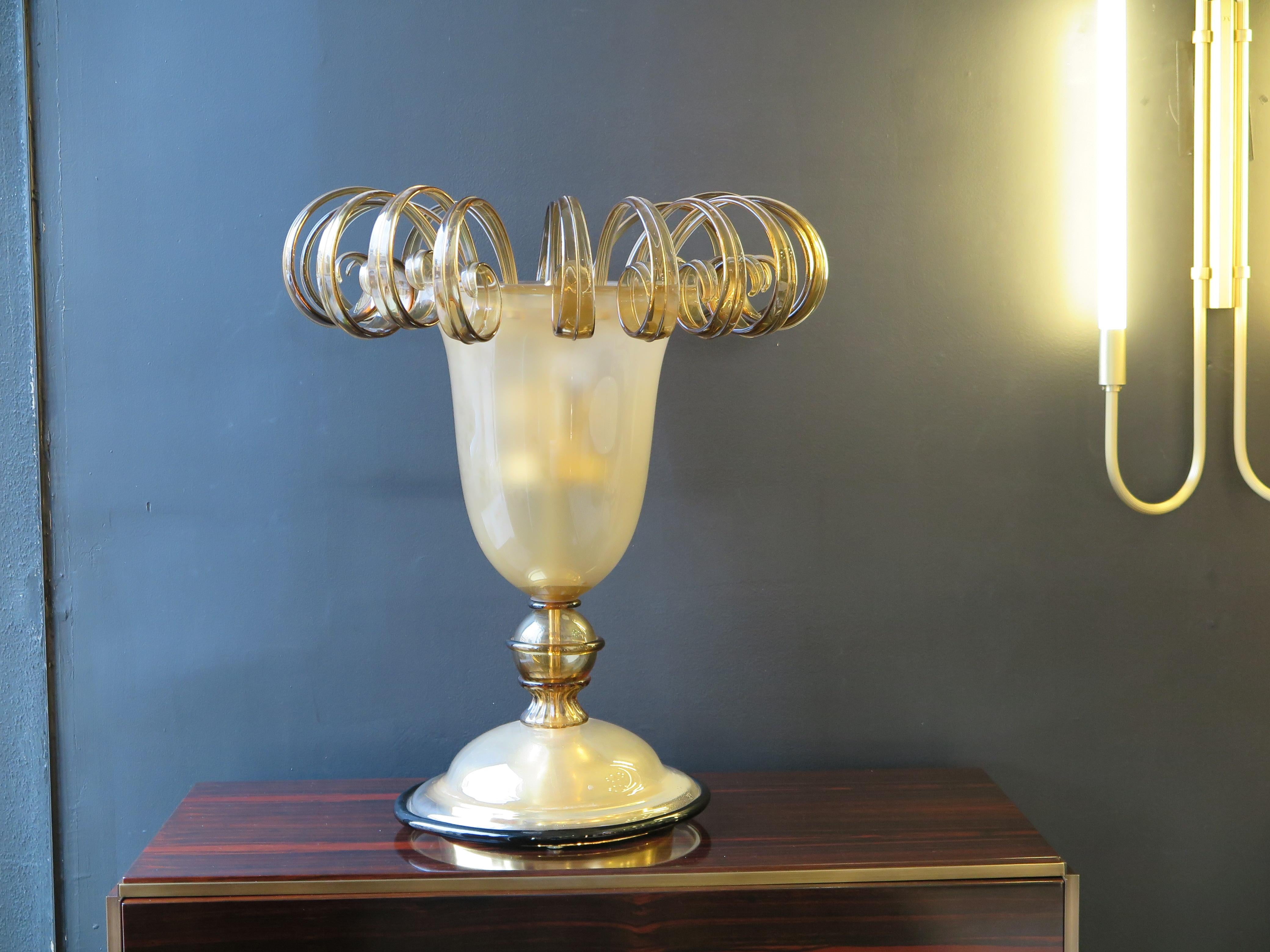 Large Italian Murano glass table lamp with 16 scroll details lining top rim in a translucent gold.  Tulip shaped lamp in gold speckled hue with black edging on base.  Three bulbs inside.
