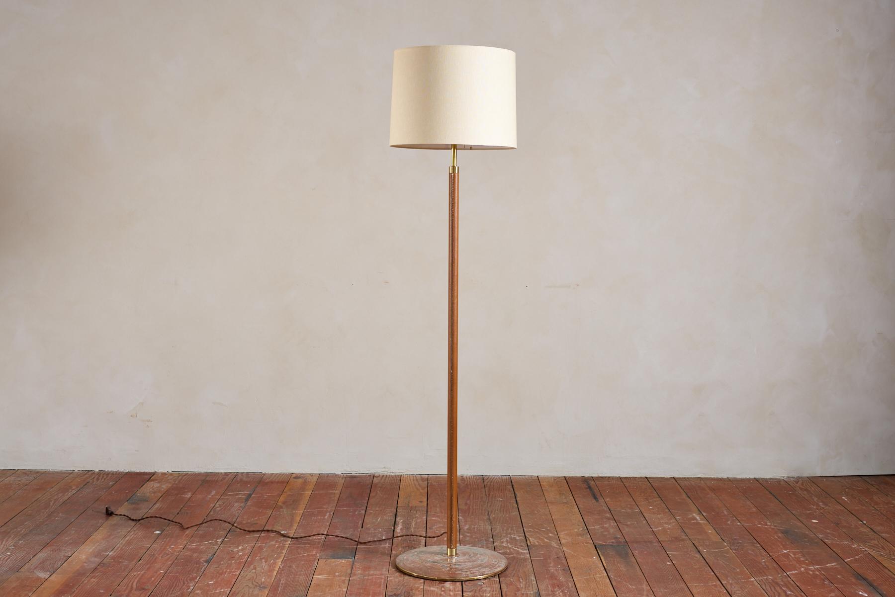 Tall Jacques Adnet style leather floor lamp 
Heavy patina'd brass circular base with caramel leather frame and new linen shade. 
Professionally rewired. 
France, 1940s