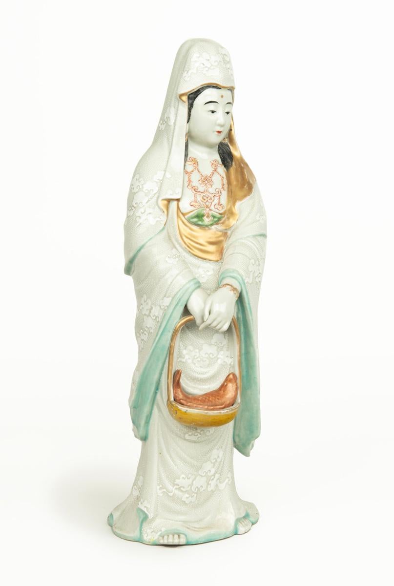 As part of our Japanese works of art collection we are delighted to offer this most charming Meiji Period 1868-1912 ceramic okimono depicting the Goddess Kannon, ( Kwannon )  the most serene figure stands clutching a basket containing a large fish,