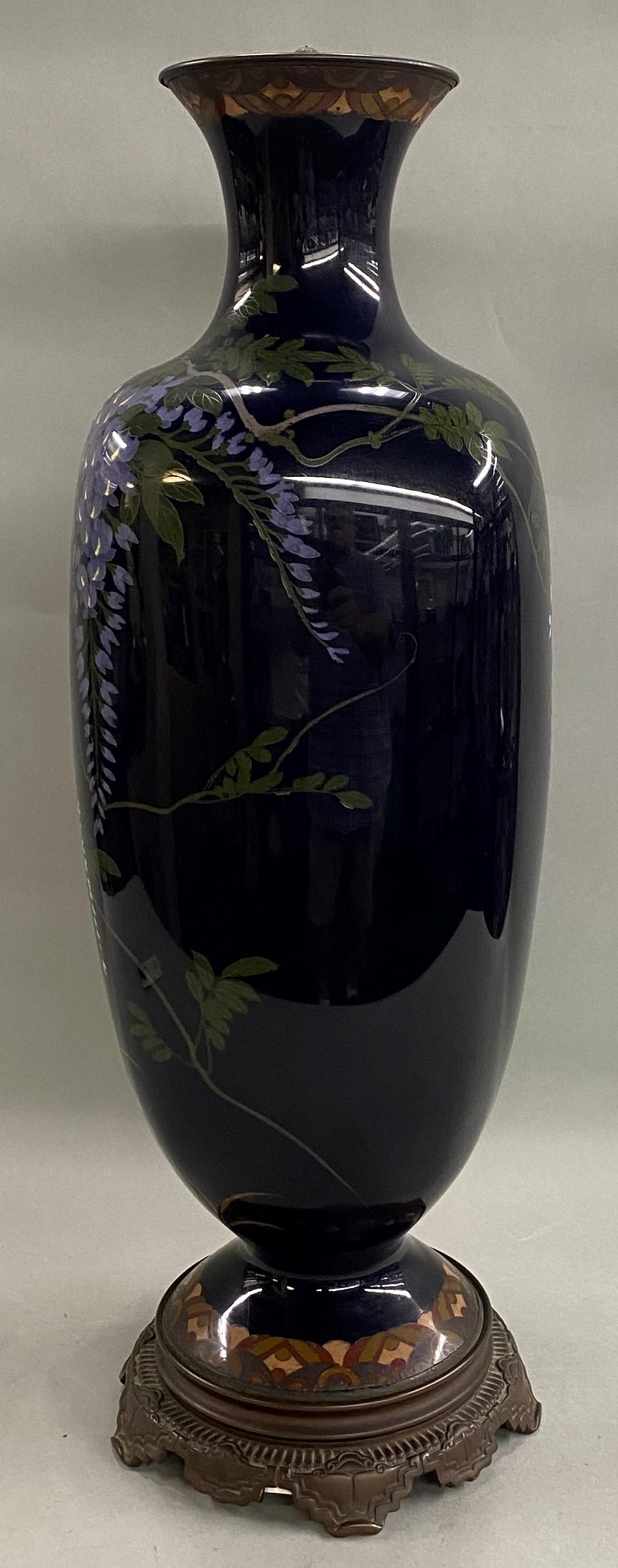 A fine large Japanese cloisonne square form vase with flared lip and round base, decorated with a large bird, wisteria, and other flowers on a deep blue background, with geometric earth tone accents around the rim and decorative brass base. The