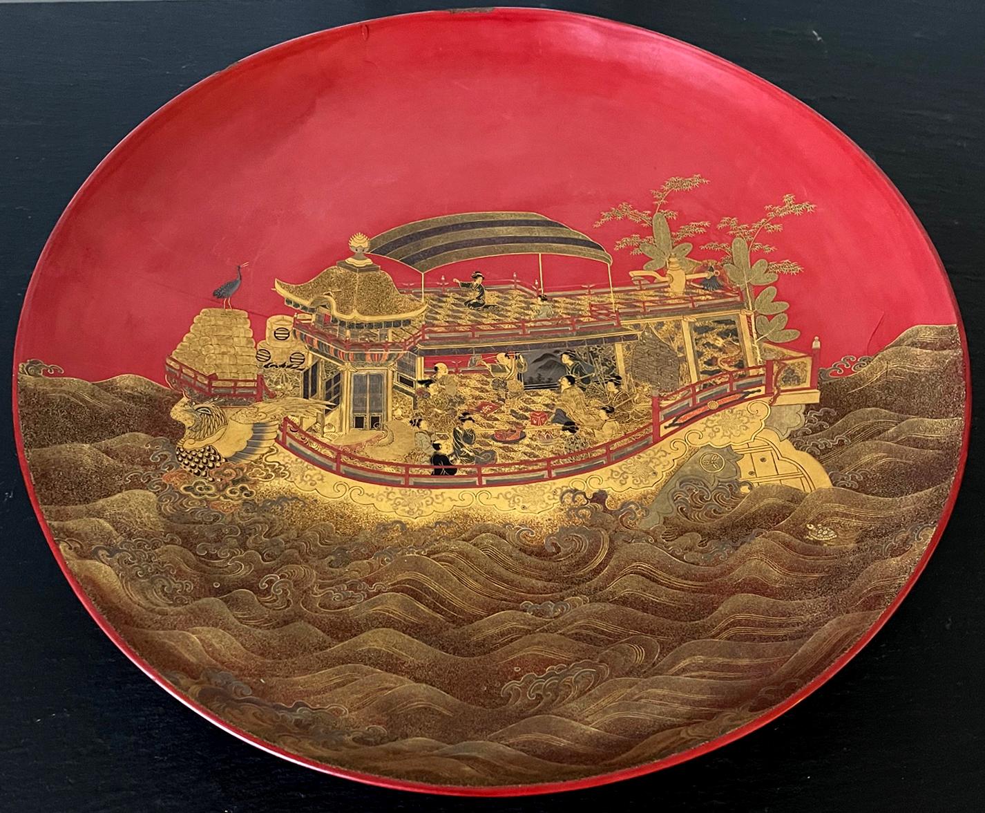 A large circular plate with a short stem base in Vermillion lacquer color. The surface was decorated with a stunning maki-e picture that depicts an elaborate boat sailing on the waves hosting a lively gathering. The boat appears to be a party vessel