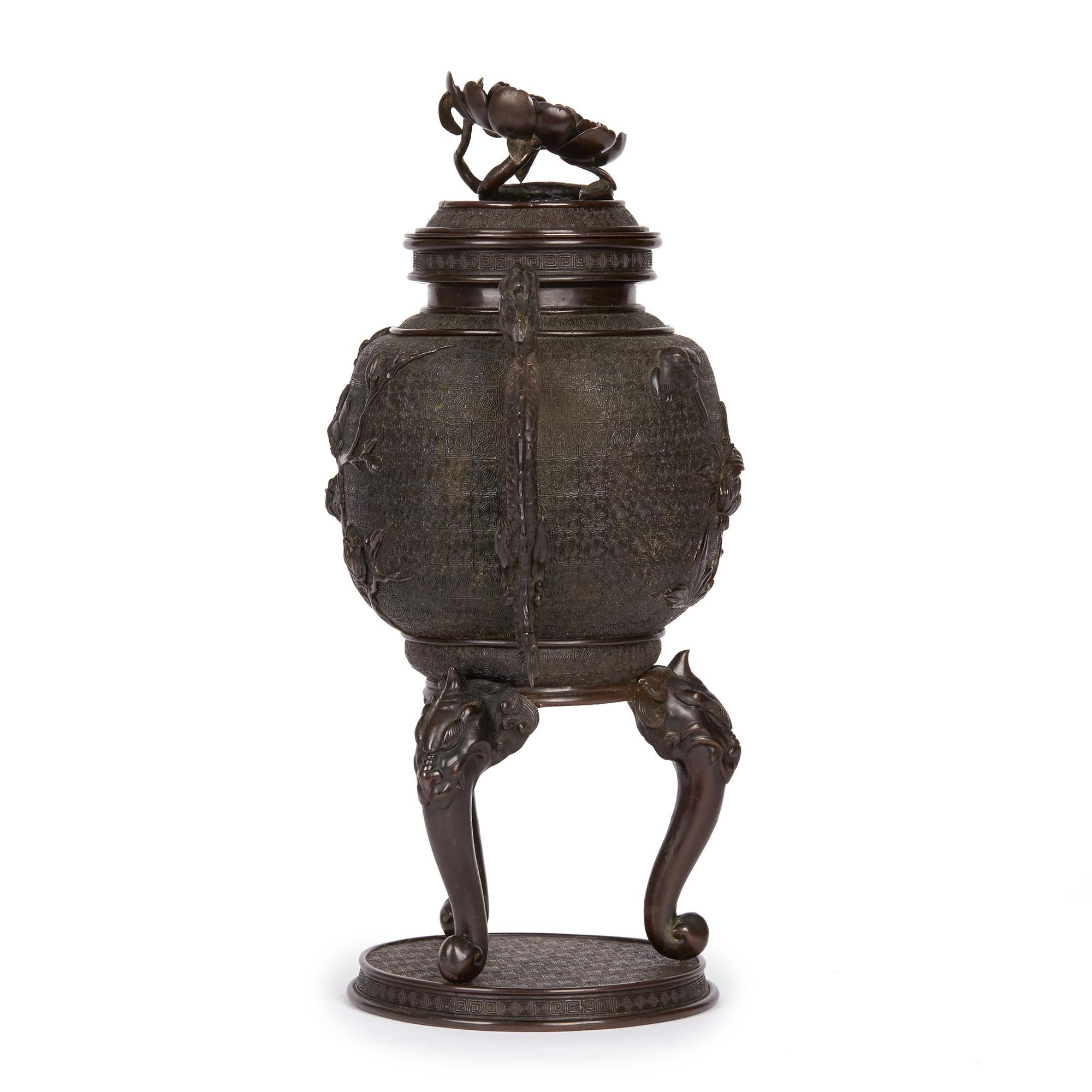 A large and stylish antique Japanese Meiji bronze lidded twin handle urn standing on a rounded plinth base with tongue and grotesque head formed legs with dragon handles and a flat cover with a large prunus flower head terminal. The body of the urn