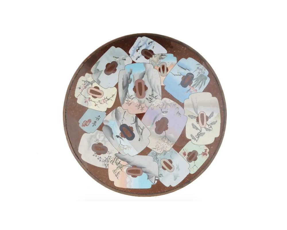 Grand chargeur ou assiette japonaise ancienne, de l'ère Meiji, en émail sur laiton. L'assiette est ornée de médaillons figuratifs polychromes représentant des paysages et des vues d'étangs, des oiseaux dans des arbres sakura, réalisés selon la