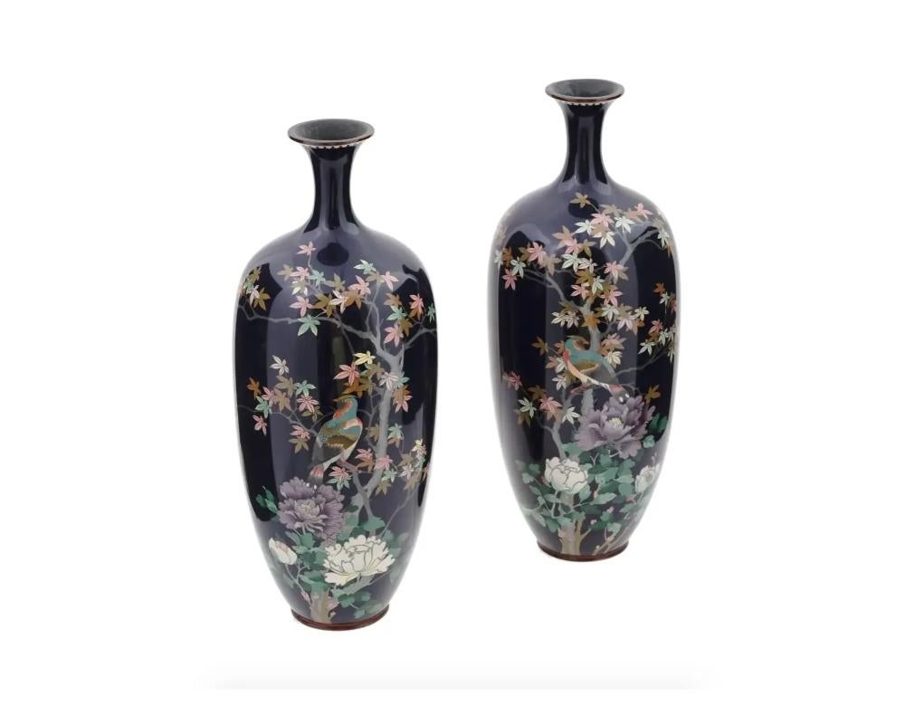 A pair of large symmetrical antique Japanese Meiji period enamel over brass vases. Each vase has an amphora shaped body and a narrow neck. Each vase is enameled with a polychrome image of a bird in blossoming flowers and trees made in the Cloisonne