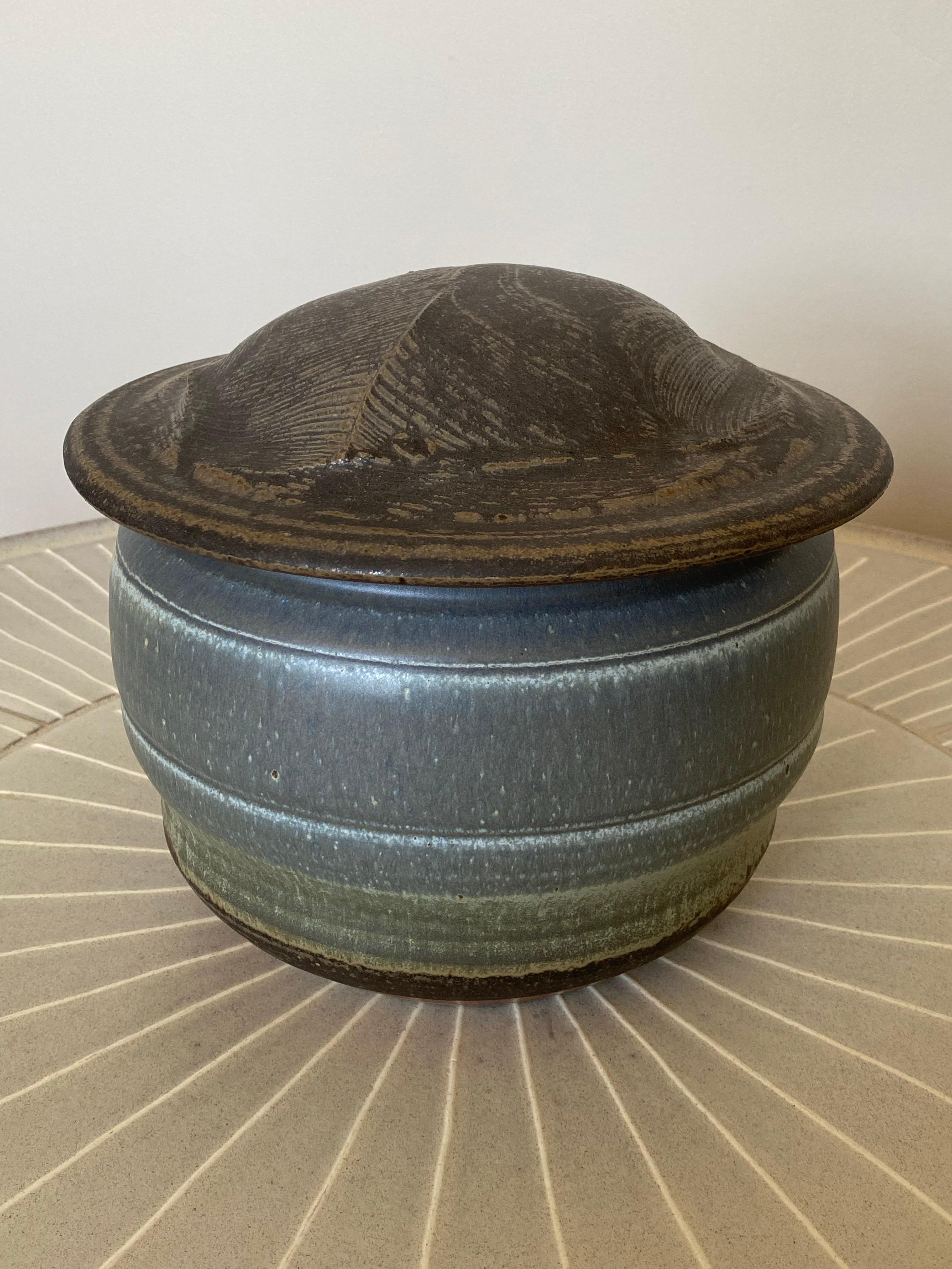 Gran jarra con tapa cortada de c1990, obra de la conocida ceramista estadounidense Karen Karnes. Esta olla presenta un vidriado en bandas de gris terroso, azul, celadón y marrón. La tapa de este excepcional tarro parece una huella dactilar. Es sin