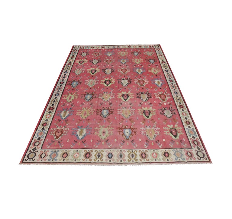 This fine wool rug is a traditional Kilim woven by hand in the early 20th century. Featuring a rich pink-red rust background with oriental motifs symmetrically woven across the open field in accents of green, brown, grey and blue. This is then
