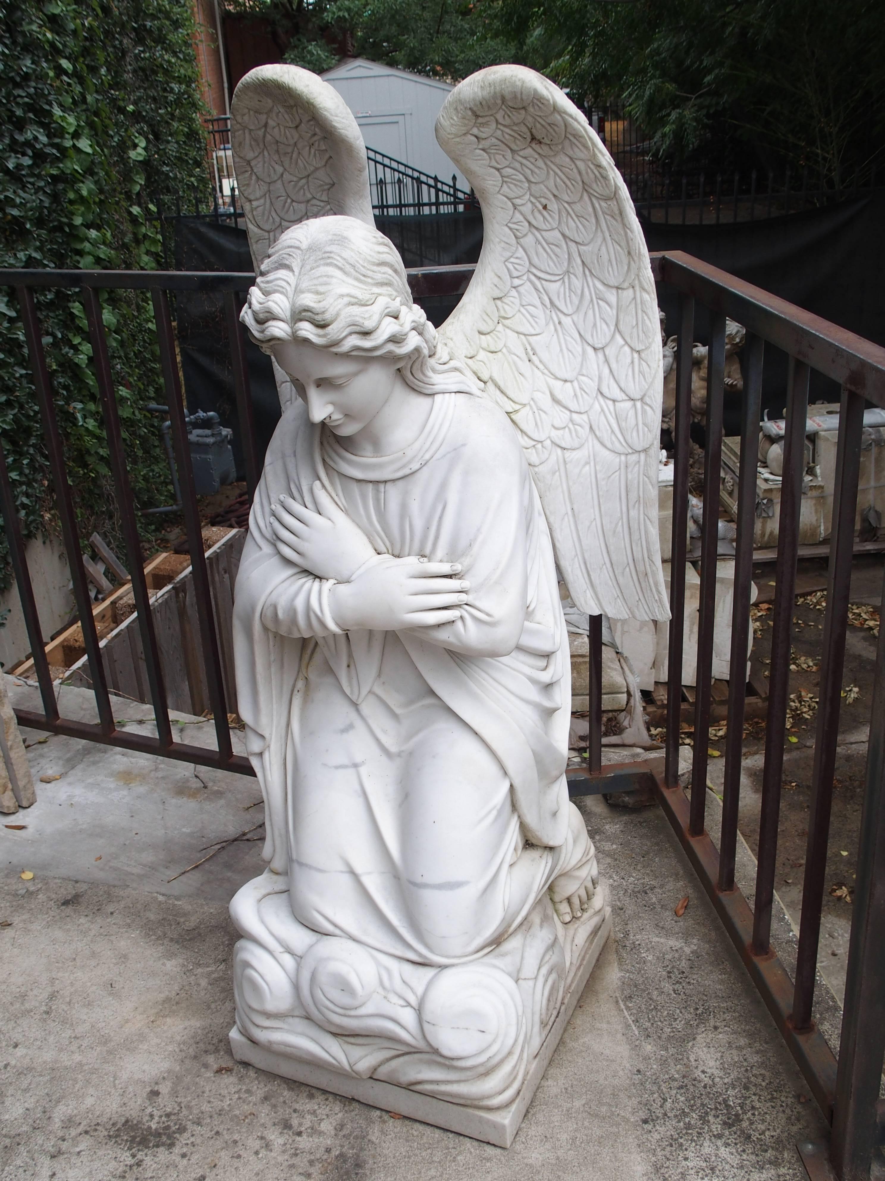 This large kneeling angel has been carved from a massive single piece of marble. The base she is kneeling on depicts stylized clouds, while she prays with her arms crossed in front of her. On her back, her large wings rise over her head in a closed
