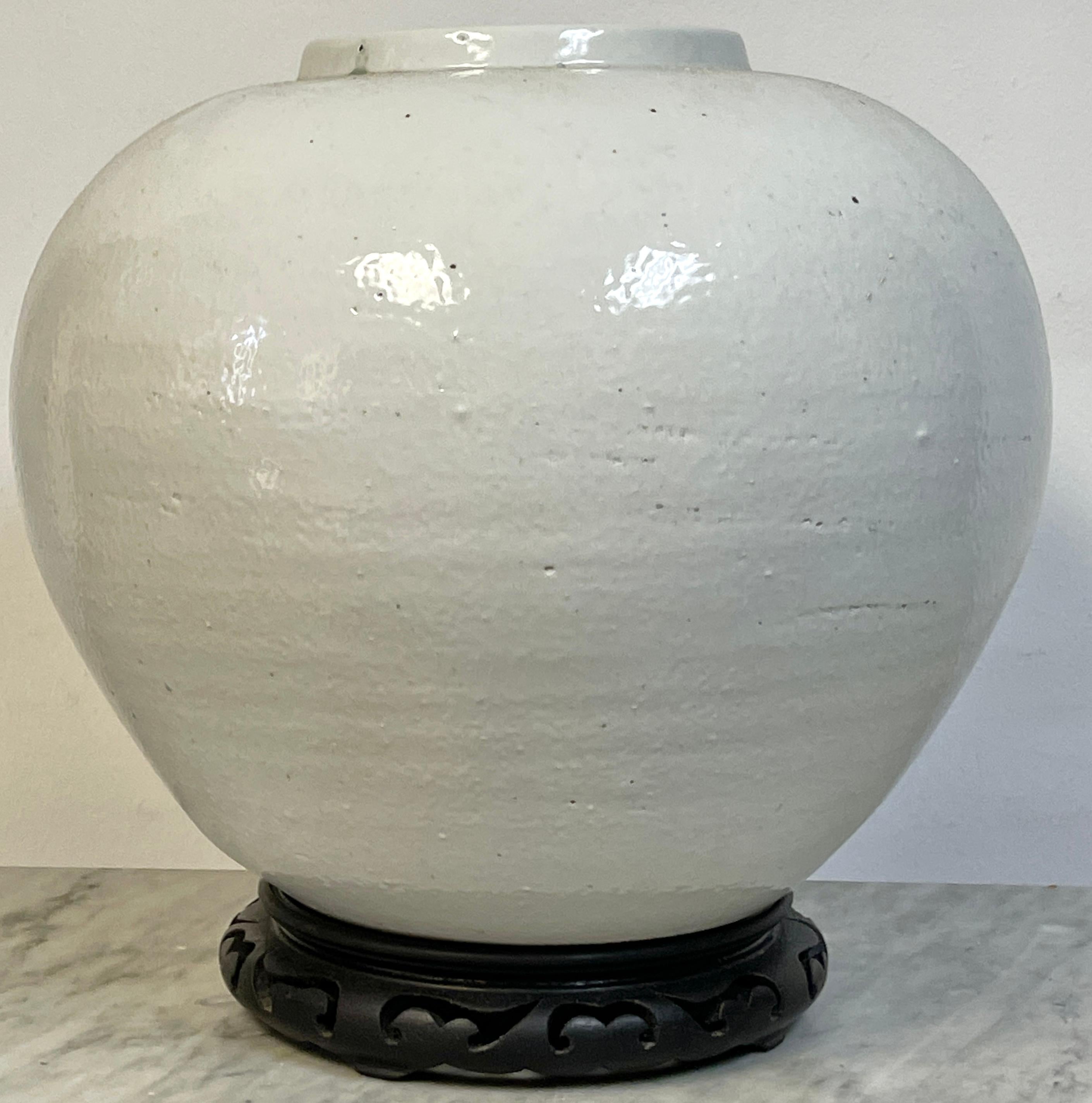 Large Korean White Porcelain Moon Jar on Carved Wood Stand, 20th Century

A striking example of Korean minimalist ceramic design, this large white porcelain moon jar embodies the refined aesthetic of traditional Joseon Dynasty whitewares. Measuring