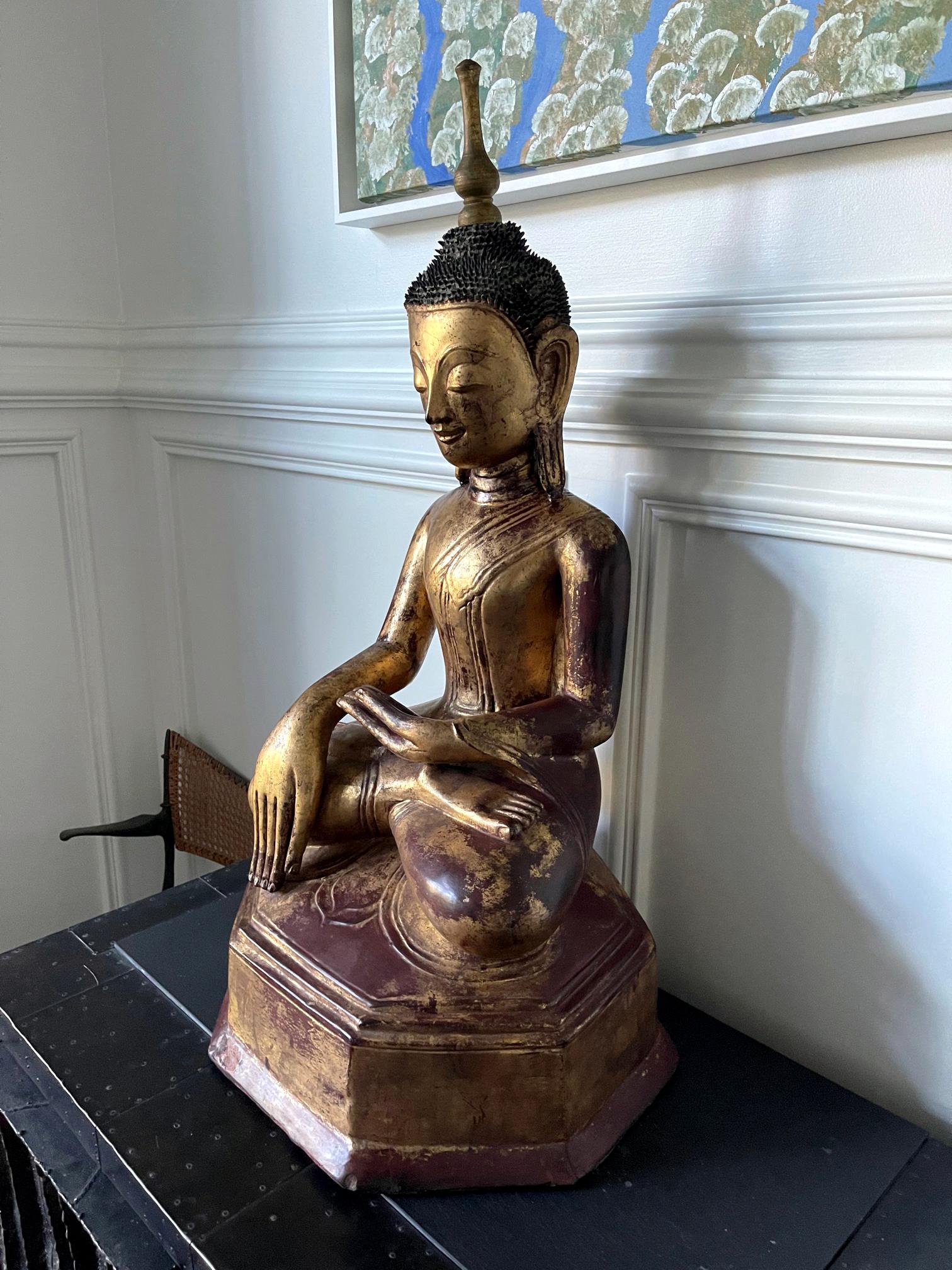 A gilt and lacquered wood Buddha statue from Southeast Asia likely Burma, circa 19th century. The Buddha is depicted as seating on a slightly tapered multifaceted plinth throne in an 