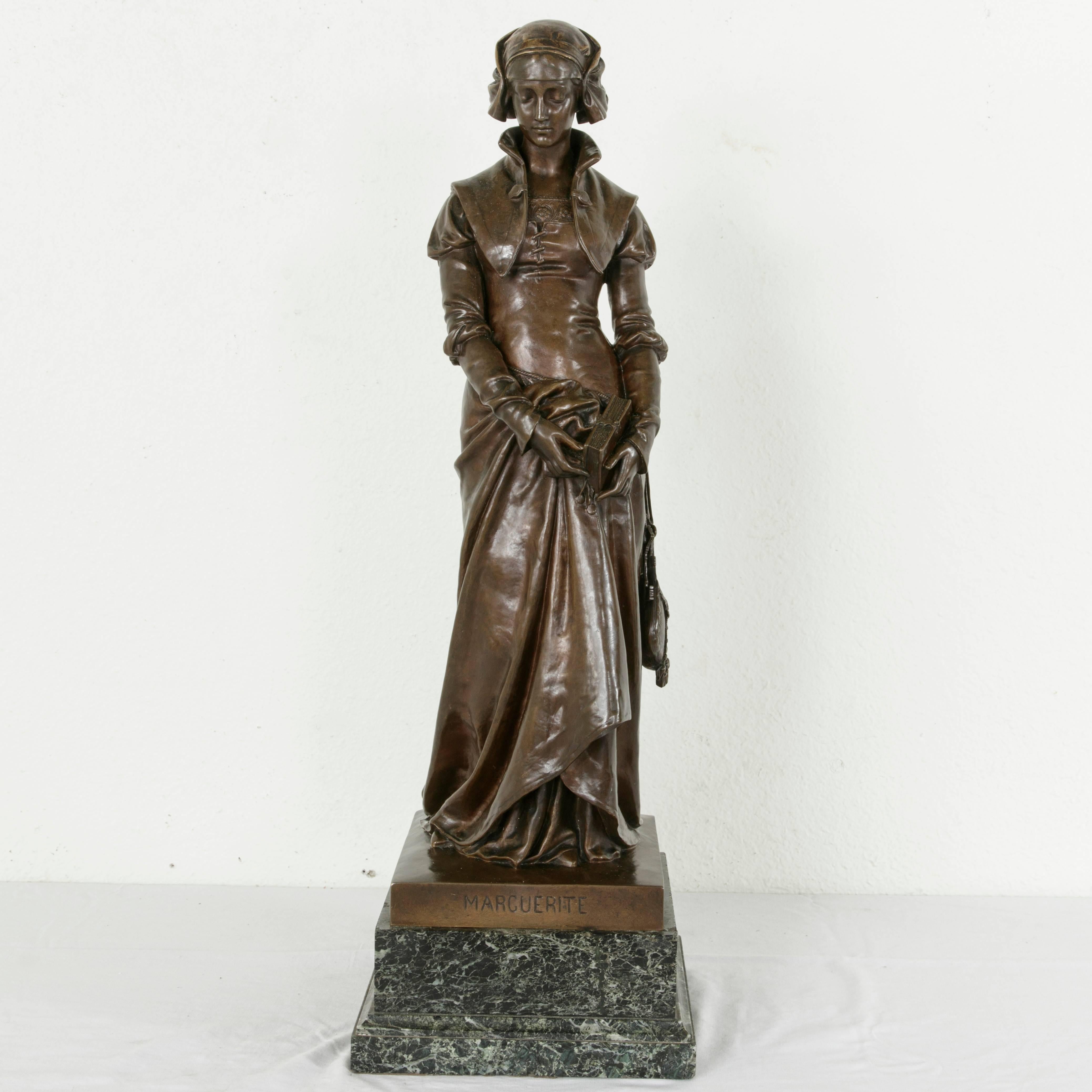 Standing at an impressive 32 inches in height, this late 19th century bronze statue on a green marble base entitled “Marguerite” features a woman with bowed head in Renaissance dress. Her hands carry a prayer book with a clasp, and rest gently at