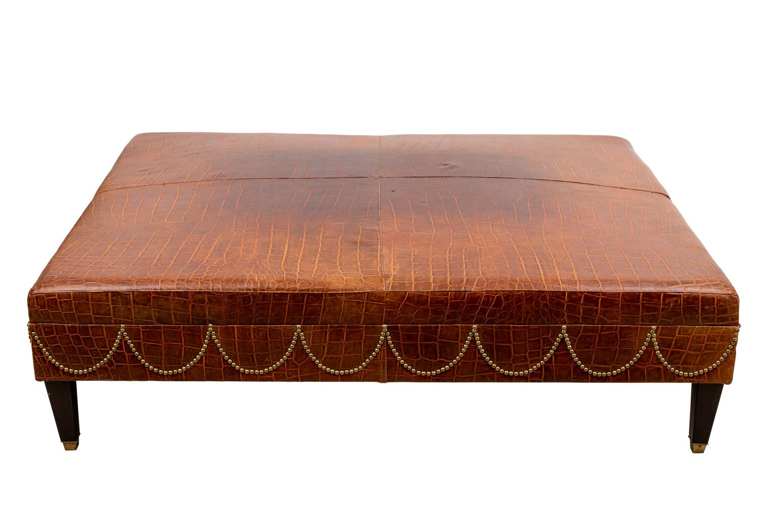 A large faux crocodile leather upholstered ottoman with scalloped nailhead trim detail on square tapered ebonized legs with brass caps.
