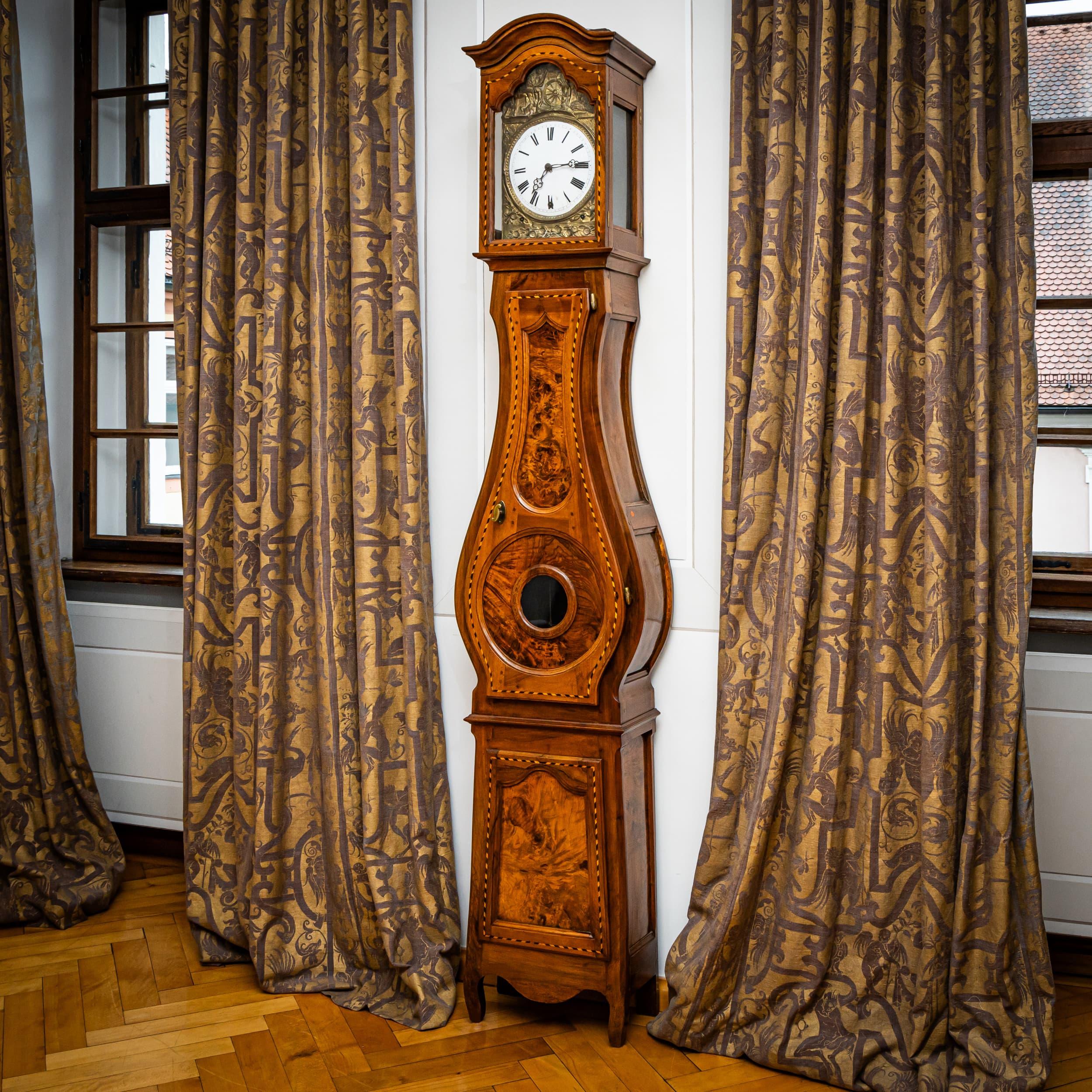Large longcase clock in walnut, decorated with fine filet inlays and a curved central section with glass panel. The clockwork is housed in the upper section with a curved cornice and features an enamel dial framed by a brass mount with rural scenes