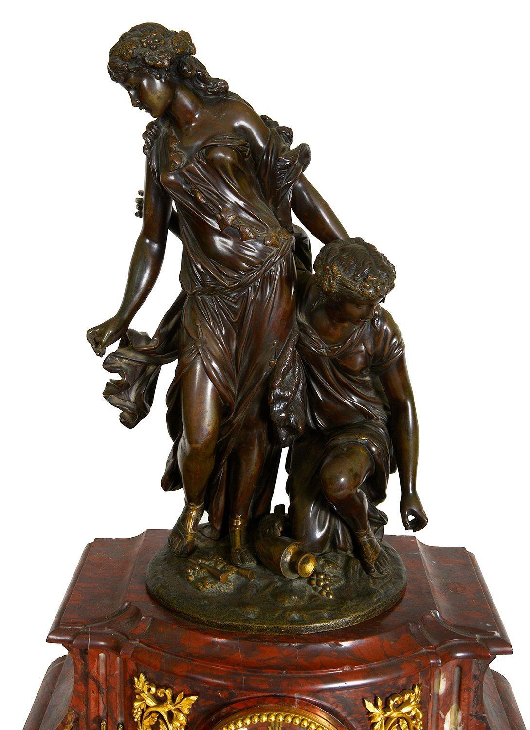 This exquisite French 19th-century clock set features a striking rouge marble base adorned with elegant gilded ormolu accents. The centerpiece showcases a detailed bronze statue of a woman and a child, capturing a moment of tenderness and grace. The