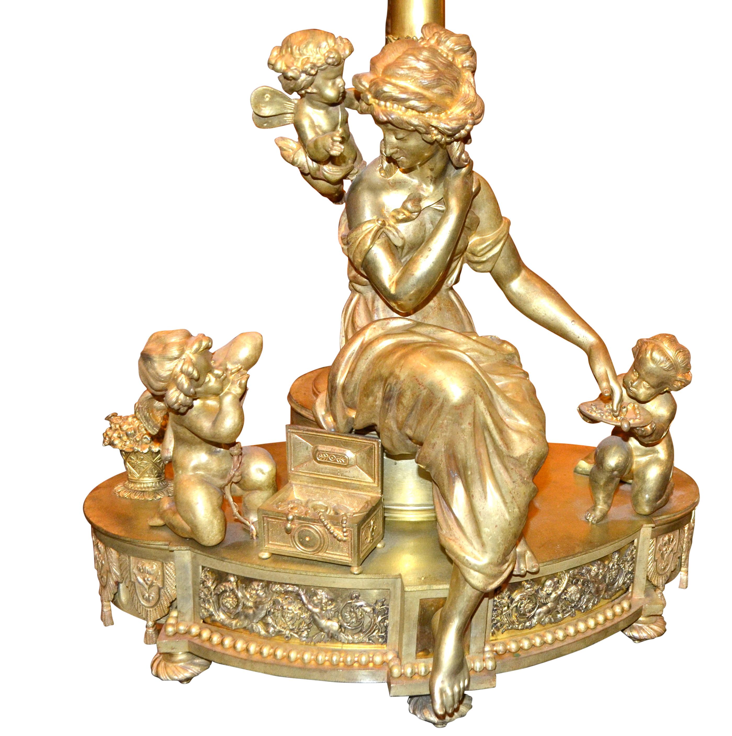 An unusual French gilt bronze candelabra, now wired as a lamp, combining Louis XVI and late Empire styles. An elegantly draped young lady sits beside her open jewellery box with three putti beside and leaning over her. Behind her rises an empire