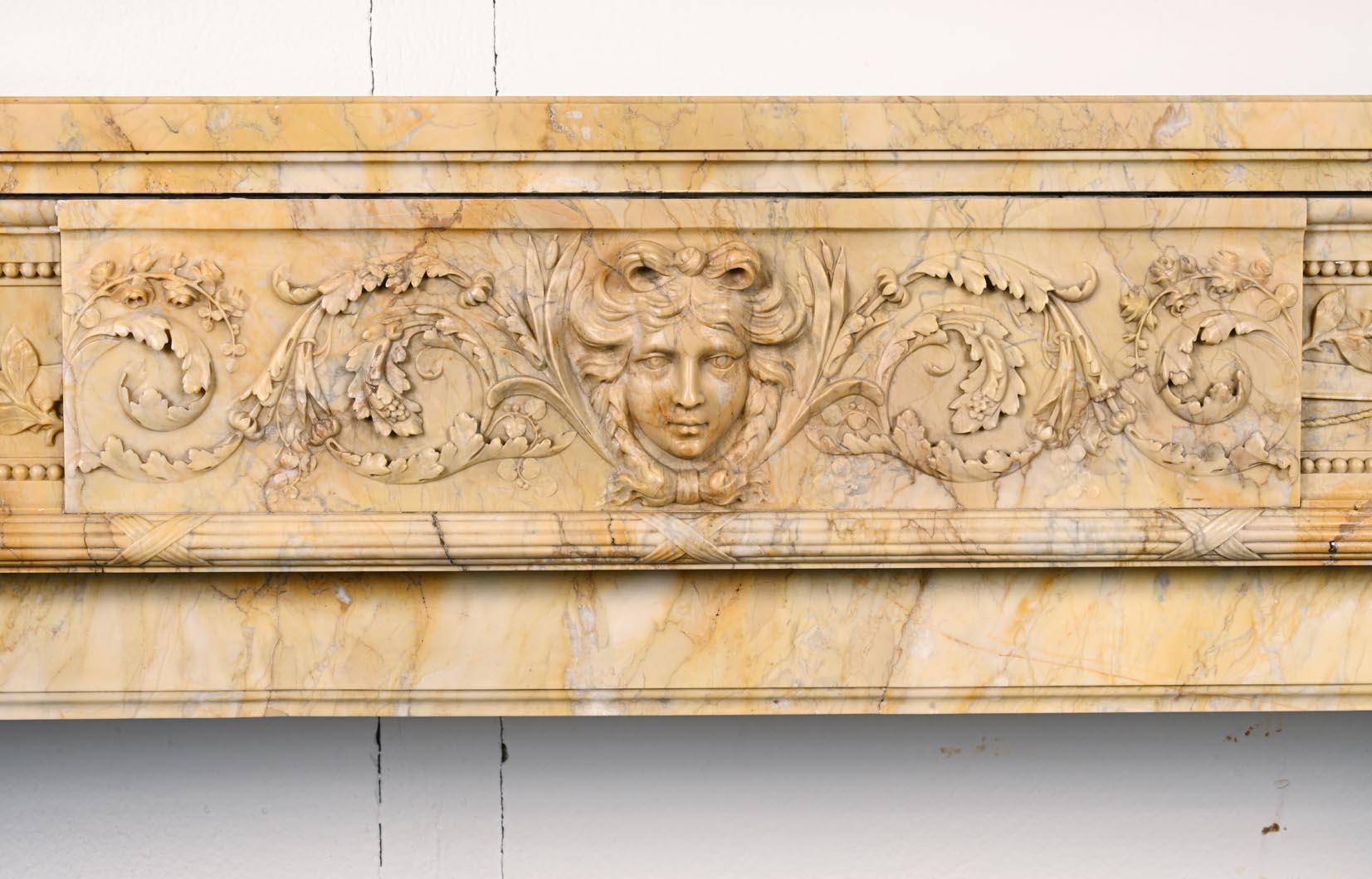 Dieser große Kaminsims im Louis-XVI-Stil wurde im 19. Jahrhundert aus gelbem Siena-Marmor geschnitzt. Den Türsturz ziert eine reiche und filigrane Flachreliefskulptur: In der Mitte ist ein Kopf des Apollo mit geflochtenem Haar zu sehen, der von