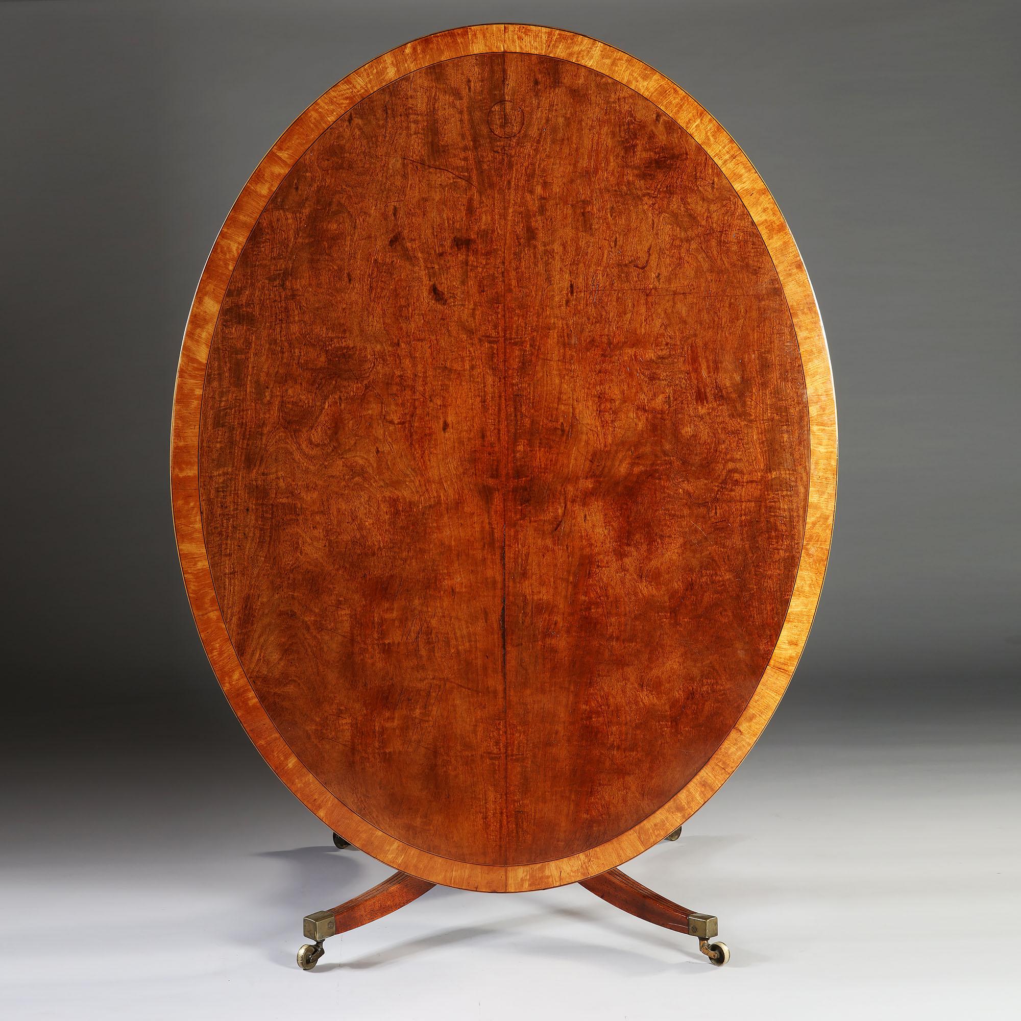 A large mahogany oval breakfast table with highly figured tilt top, including an inlaid border, resting on four fluted legs terminating in brass castors.