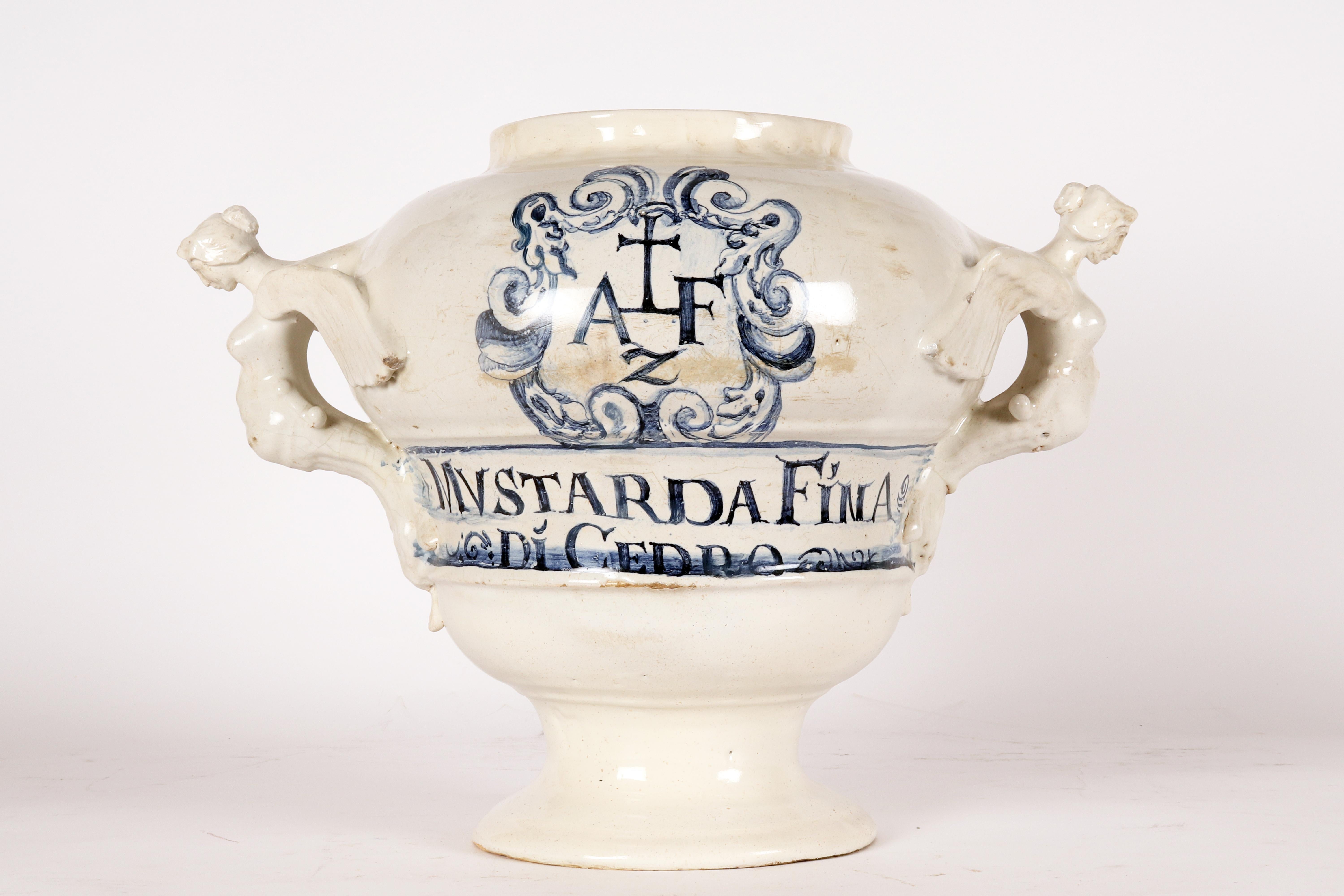 Large majolica Albarello with a flared central body with curvatures at shoulder height, wide distinct foot (8,75’’), flared and moulded. Wide mouth, strongly projecting harpy-shaped handles with lower attachment with drape and tassels. On the