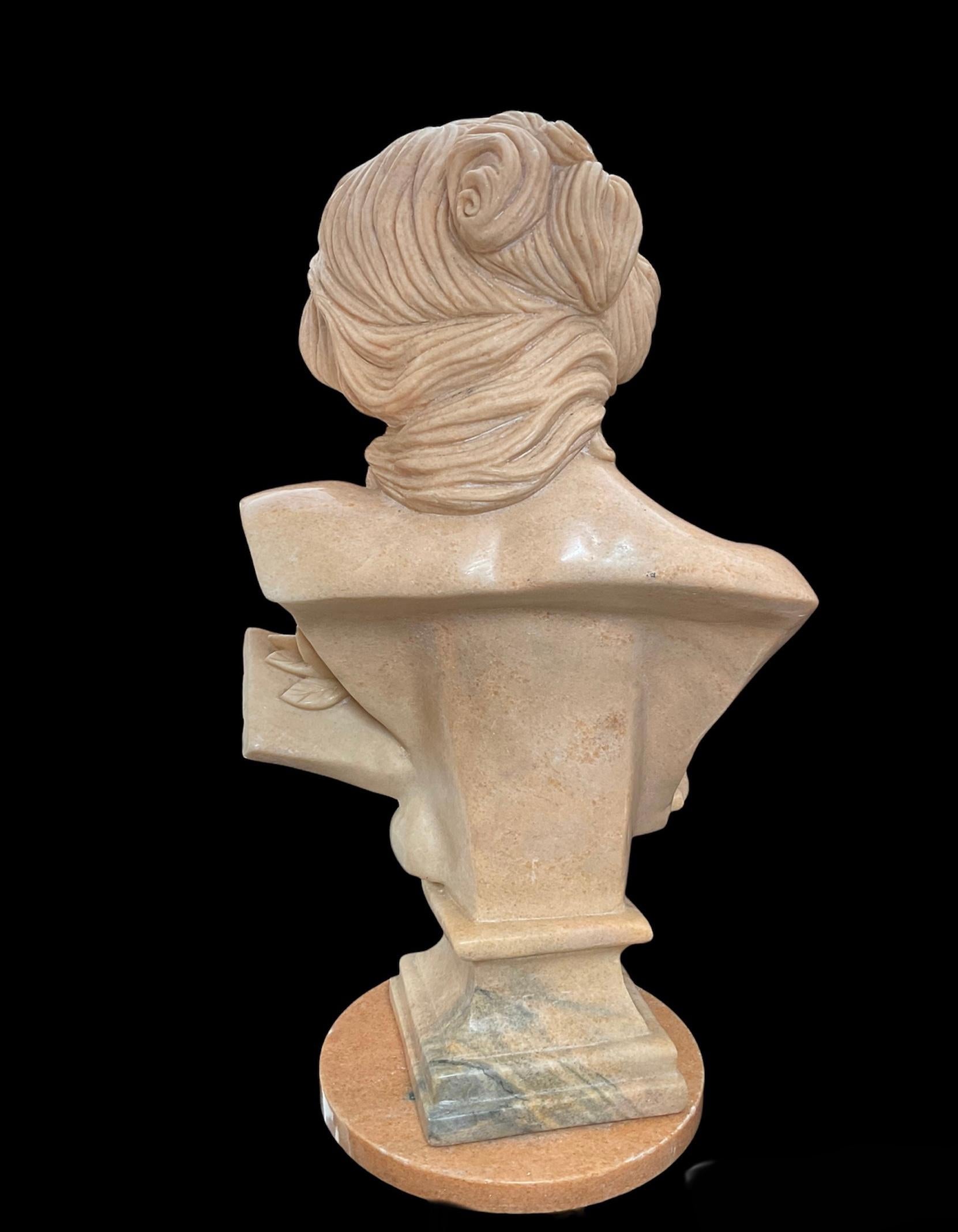 This a large rose beige marble bust of a modern music goddess. She has abundant wavy hair arranged to the side. Her head is tilted to the same side of the hair while she is looking down with a sweet gaze. Her chest is semi-covered by a robe. One of