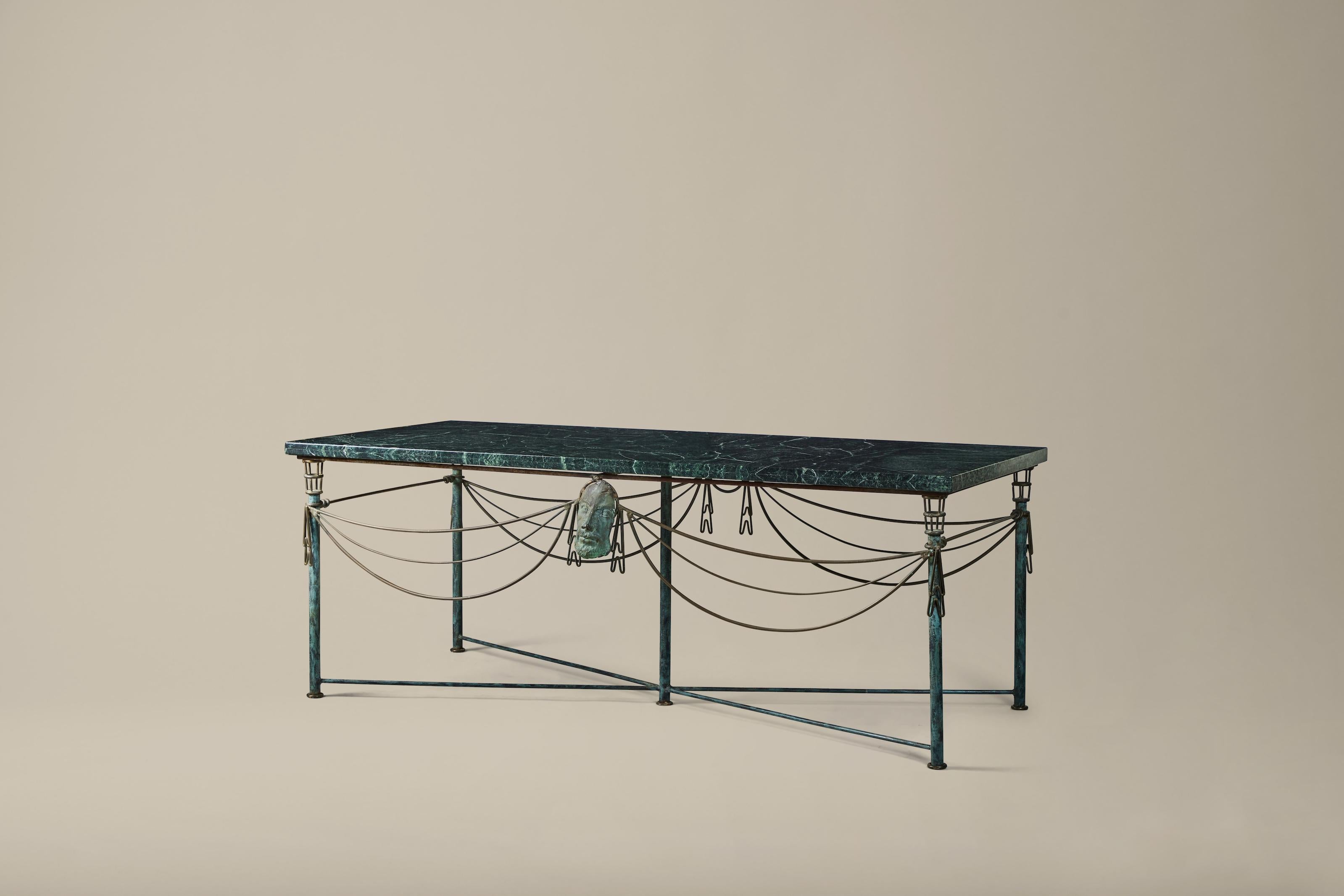 This patinated metal and marble table was designed by Nicaraguan artist Mario Villa (1953-2021). The exaggerated swag and Grecco-Roman mask apron are characteristic of Villa's work. The table features a verdigris marble top, cylindrical legs,