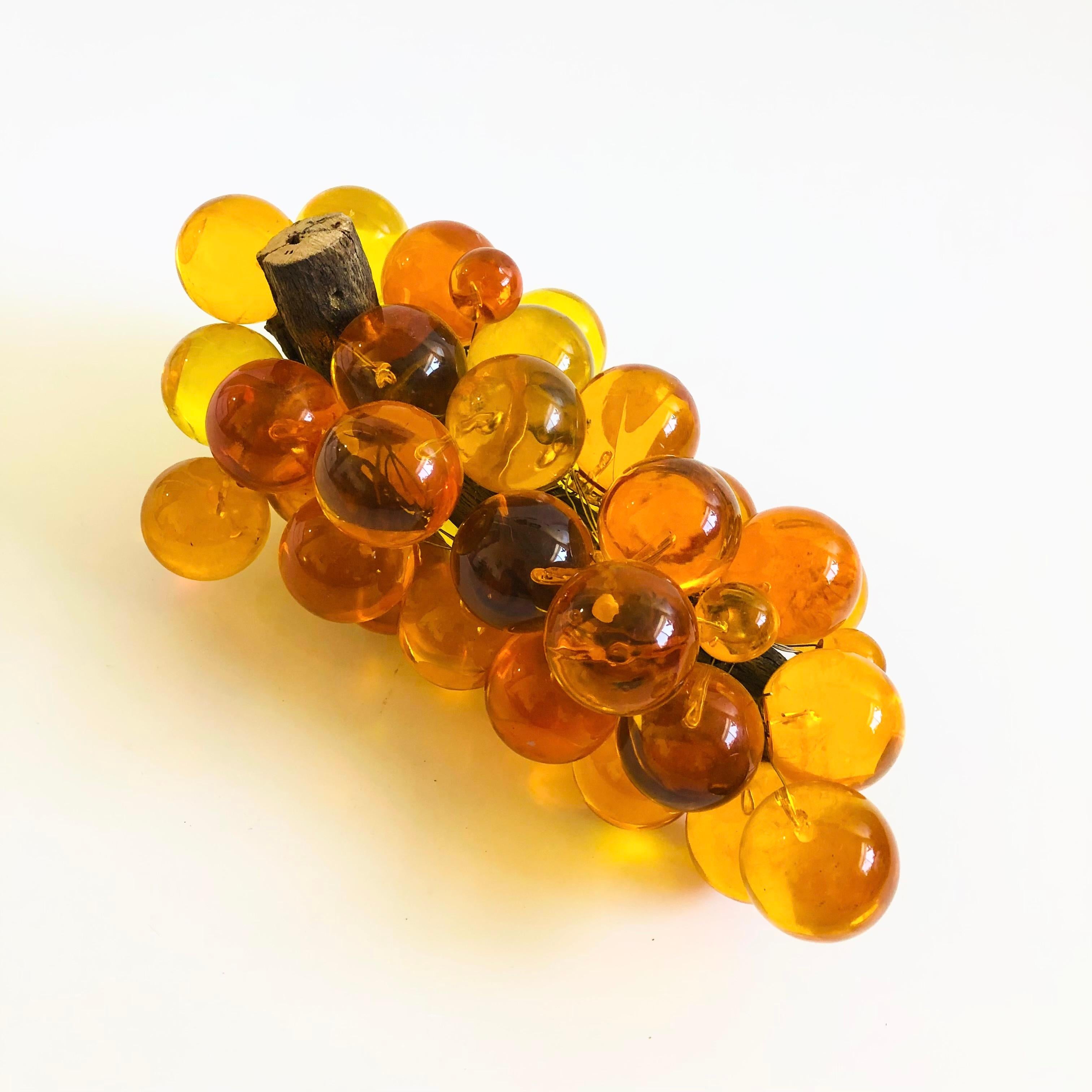 A mid century lucite grape cluster. Spherical lucite grapes are held together by metal wires. Fastened on a natural wood stem. Beautiful variation in the amber color of the grapes with a few small grapes tucked in between the large grapes. A great