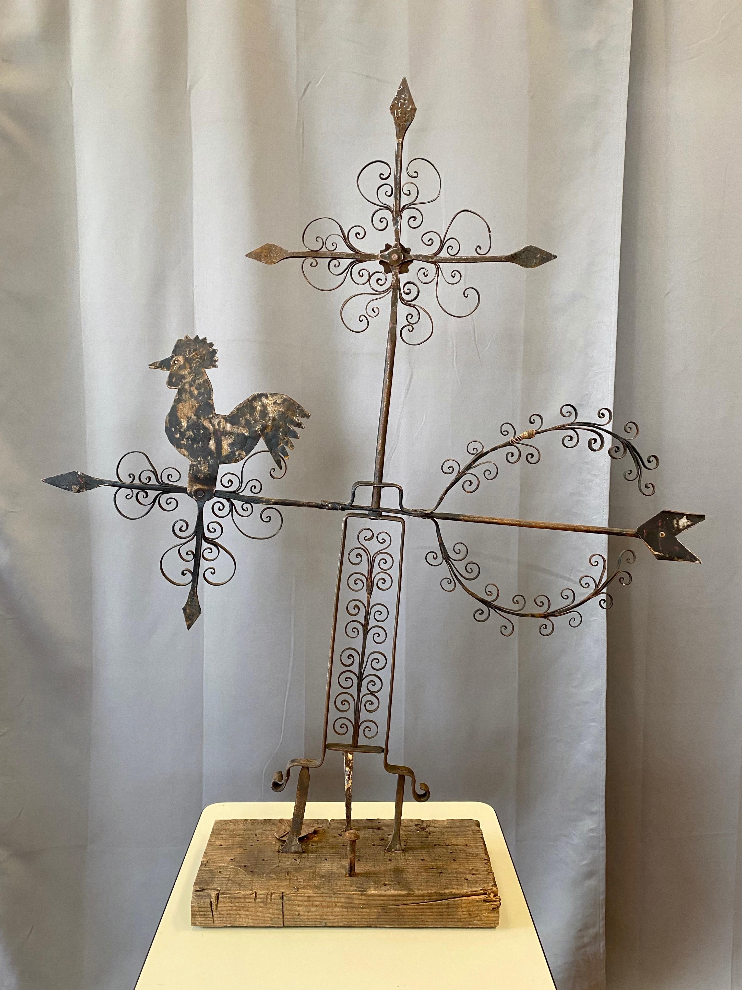 A large circa 1940 Mexican Folk Art wrought iron rooster weathervane presented on a rustic reclaimed wood base.

Features a sheet metal rooster perched upon a directional arrow, with wrought iron scroll work on delightful display throughout. Topped