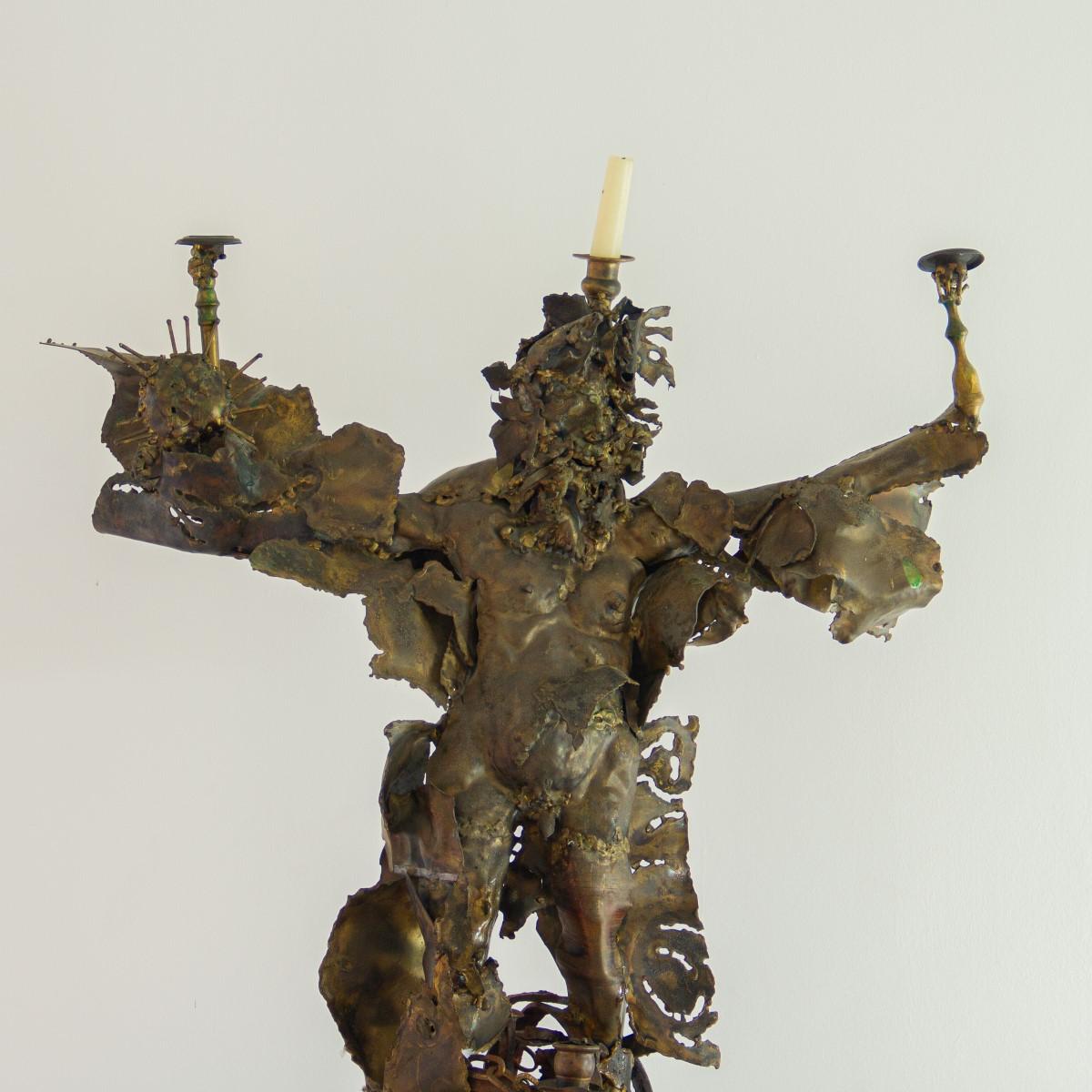 A large Mexican Brutalist, torch cut metal sculpture featuring a bearded figure balancing on an armillary sphere, which is set on an ebonized wooden base. The sculpture has four candleholders, one in each hand, one on his head and another coming out