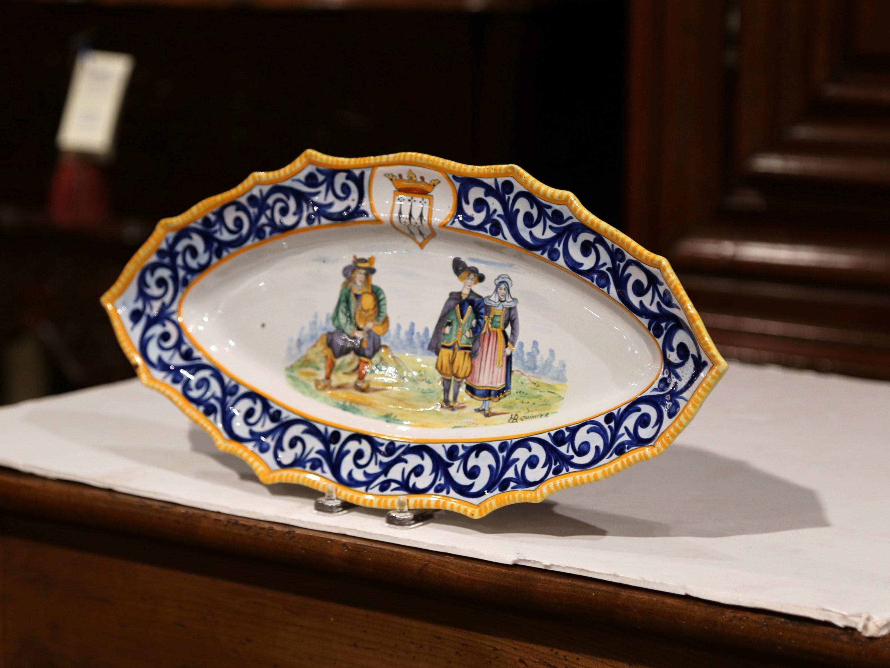 Decorate a wall or a shelf with this colorful, antique platter. Created in Brittany, France circa 1960, the hand painted ceramic plate depicts a Breton couple in traditional costumes and a seated man playing the bagpipe. The oval, porcelain platter