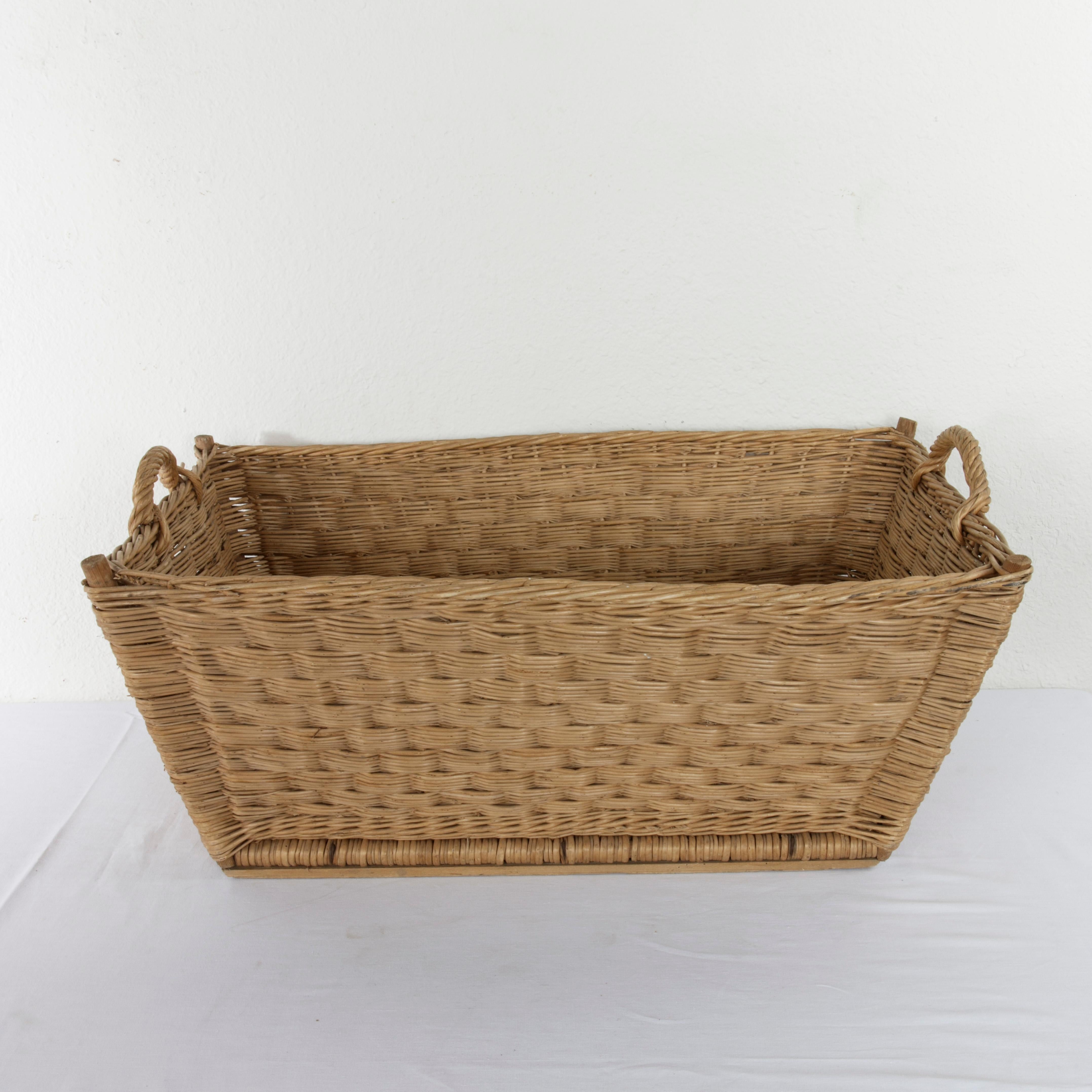 Originally used for carrying laundry to be hung outside, this large handwoven rattan basket from the mid-20th century features a tapered shape around its wooden frame. A twisted woven handle at each end allows for easier carrying. Its base measures