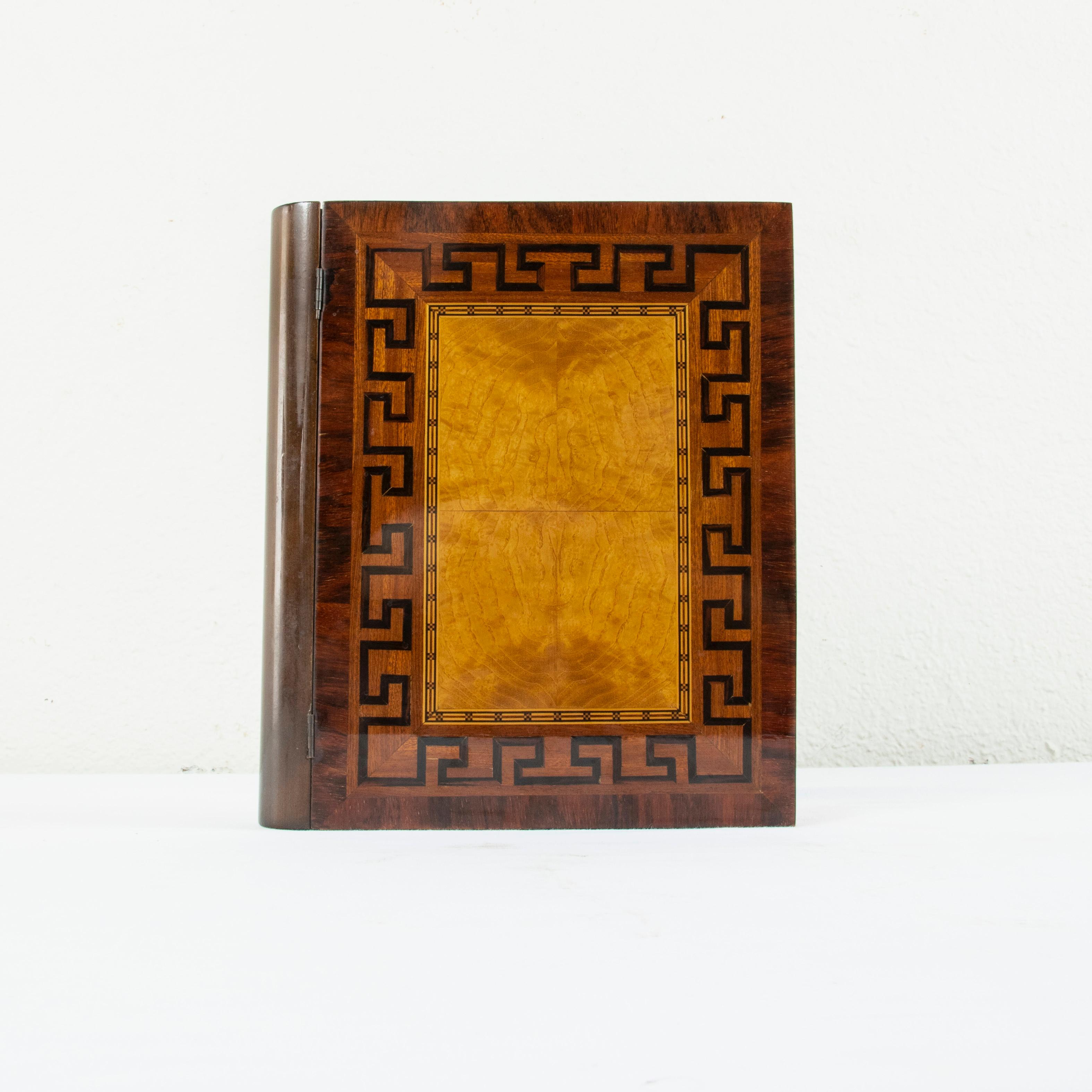 This mid-20th century French mahogany and maple marquetry box takes the shape of a large book. The lid of the box features a central square of book matched lemon wood surrounded by fine lines of lemon wood, and ebonized pear wood in a geometric