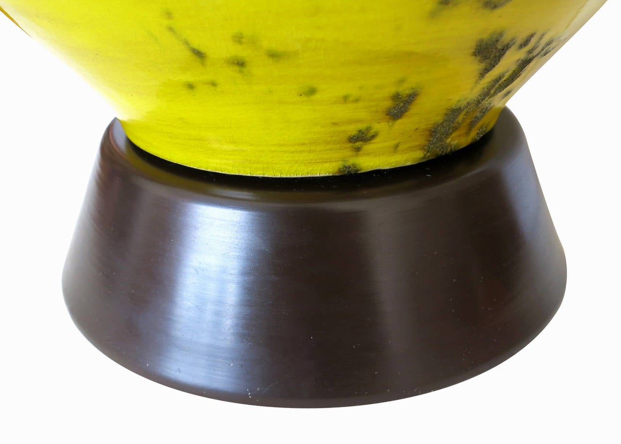 Made circa 1960, the ceramic lamp has a brown and yellow glaze finish that is spotted with textural areas. The lamp is of a unique hourglass shape, sits on a dark stained base, and has brass hardware.

