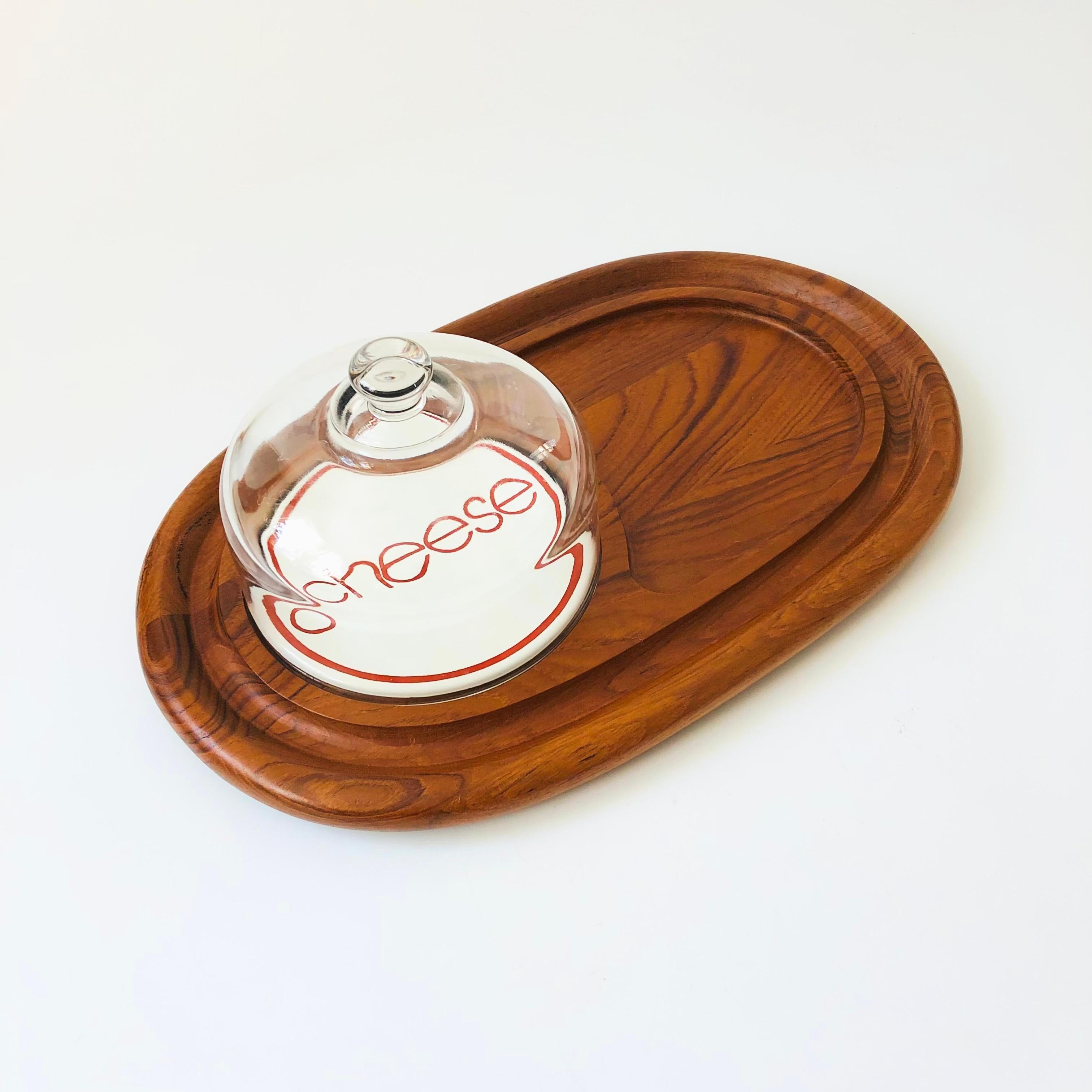 A mid century teak cheese and cracker serving tray. Nice long oval shape. Features a glass cloche on one side over and enameled plate with the words 