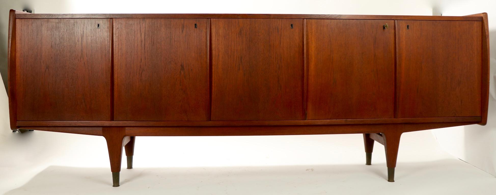Exceptional Danish modern sideboard credenza of solid teak with four lockable doors, sculpted legs, and brass feet. The doors open to reveal shelved space and spaces with drawers. The case has sculpted sides which extend beyond the doors and top to