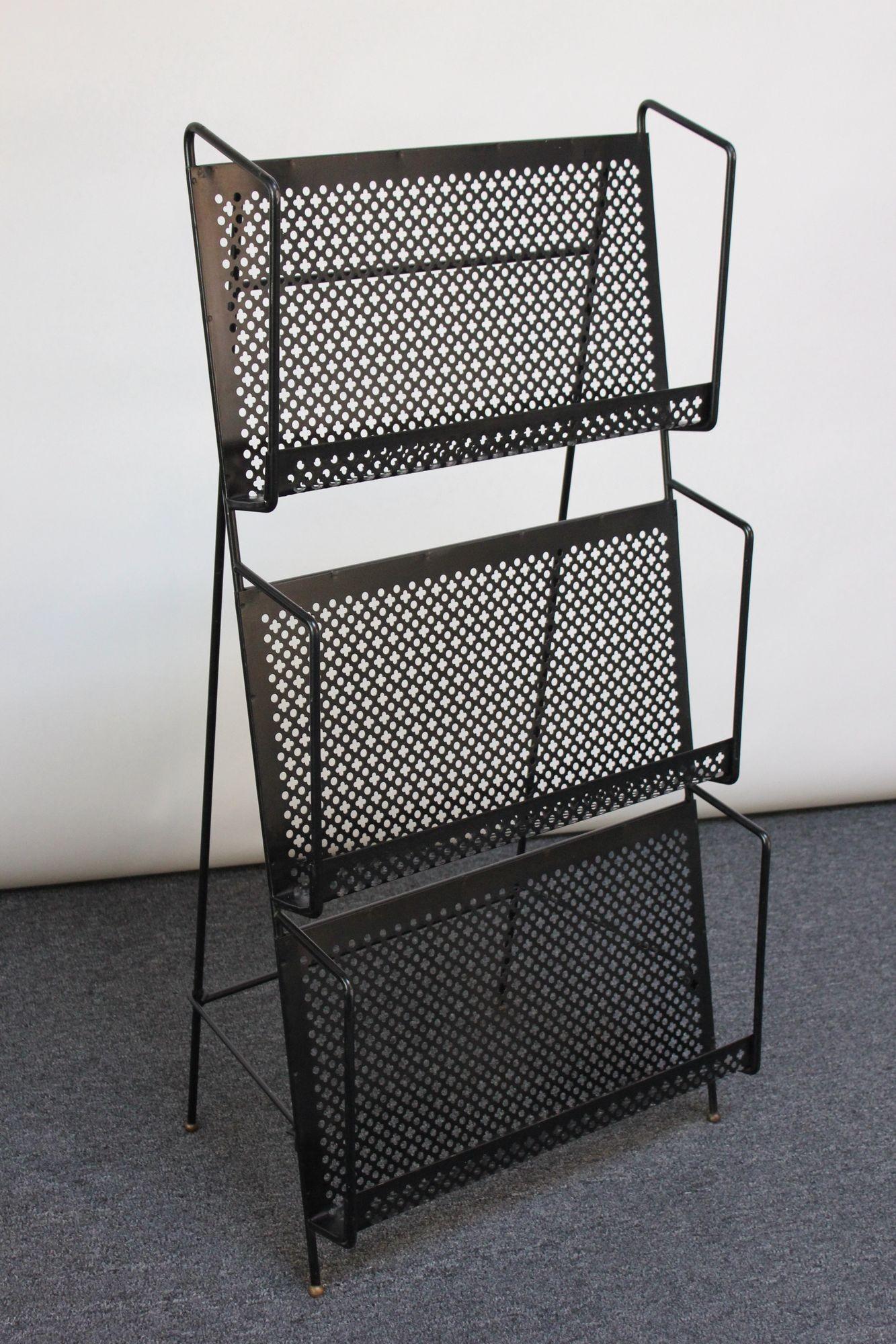 Large, free-standing three-tier magazine rack in black enameled metal with perforated cross and circle motif, all supported by gold, orb feet (ca. 1950s, USA). Each tier is 2.75