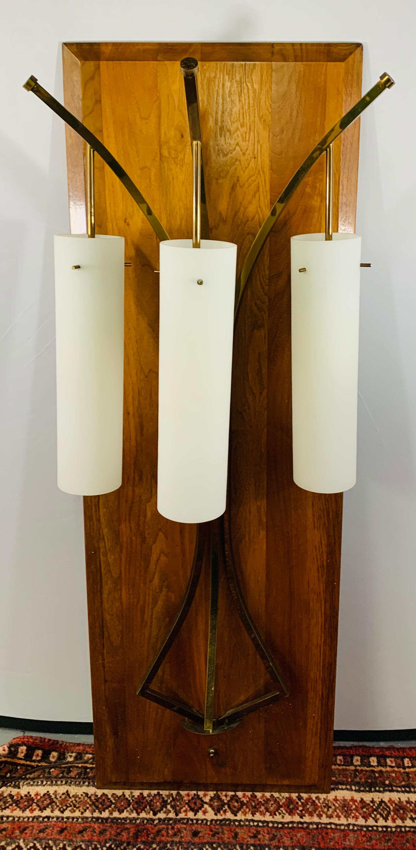 A large Mid-Century Modern wall fixture with brass frame. The fixture has three lights, each in a cylindric shape and made of milk glass. The lights are attached to a rectangular wooden panel. There is one switch that controls the three light and