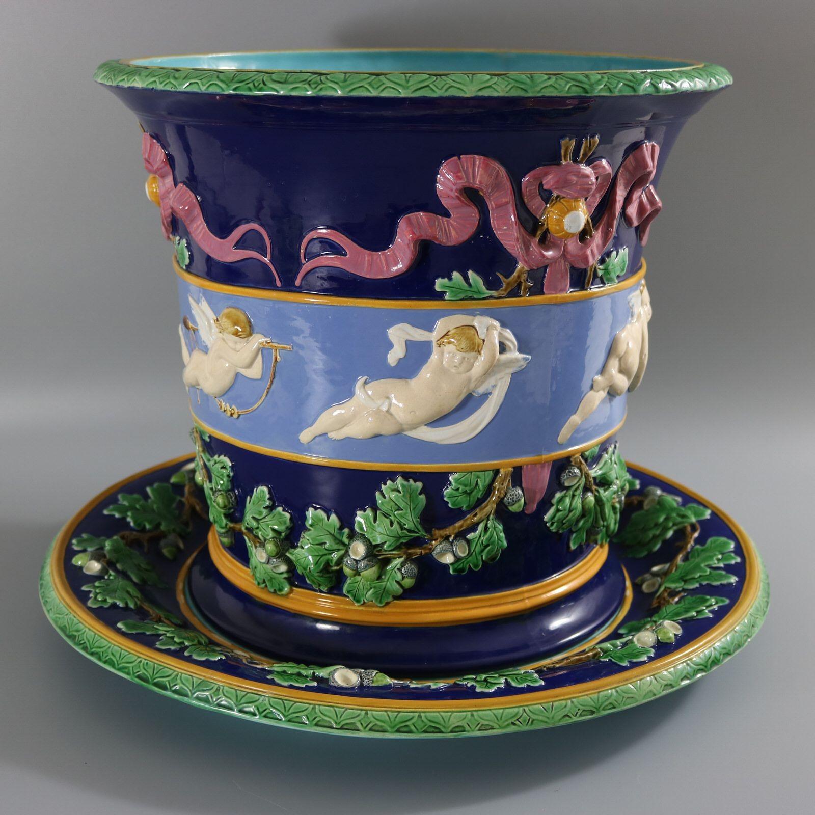 Minton Majolica jardinière and stand which features cupids in flight in various poses, around the central band. Ribbons above, oak leaves and acorns below. Colouration: cobalt blue, lavender blue, green, are predominant. The piece bears maker's