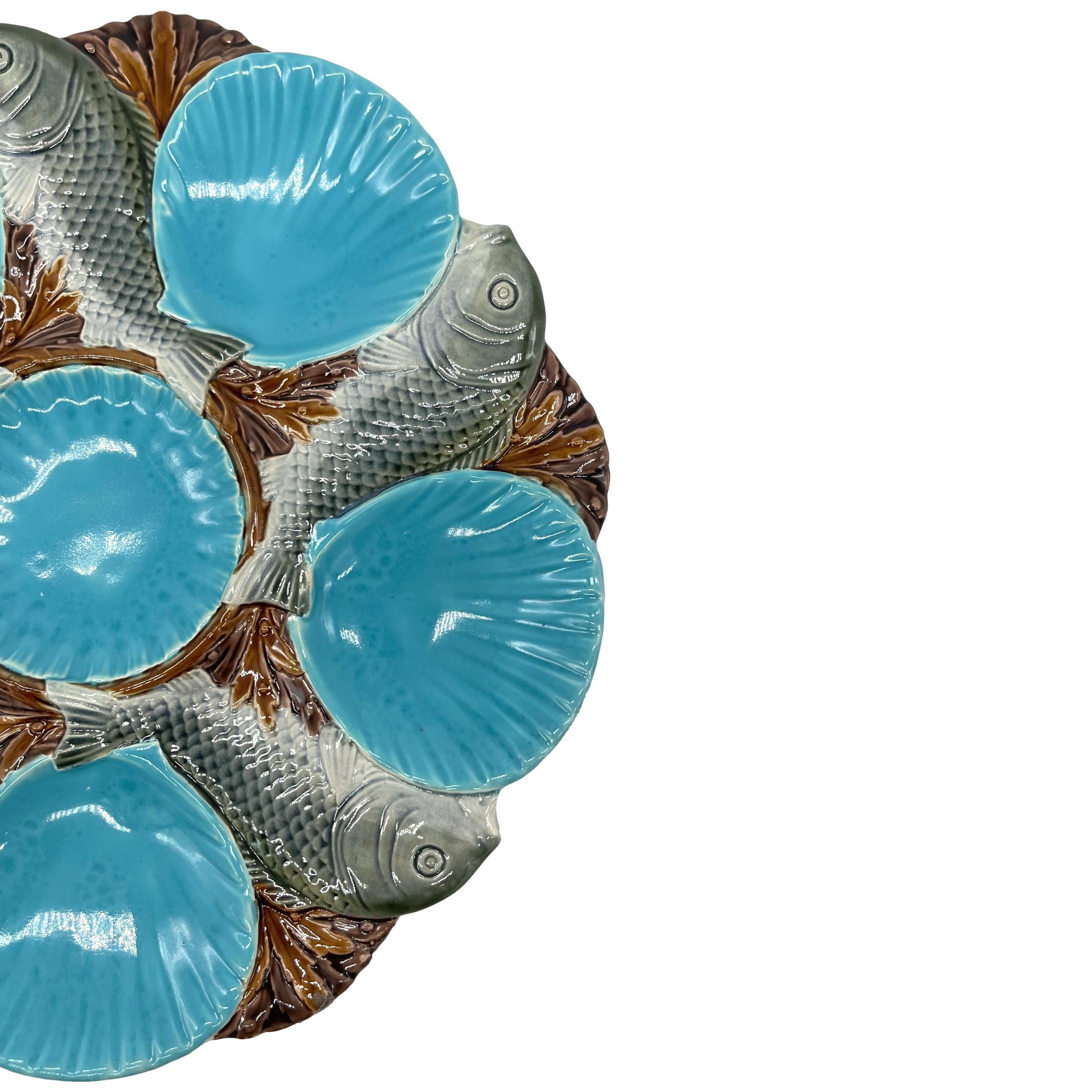 A Minton Majolica Oyster Plate, molded with a central scalloped well within five shell-form oyster wells, all glazed in turquoise blue and separated by spiraling fish naturalistically glazed on a bed of seaweed, with impressed marks to reverse,