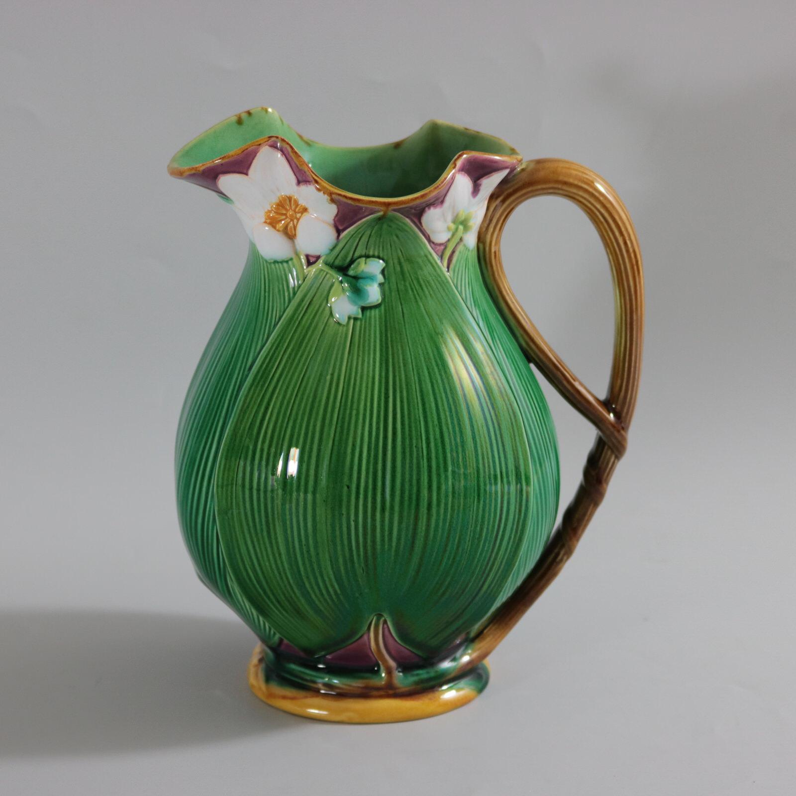 Minton Majolica jug/pitcher which features flowering lilies and lily pads. Colouration: green, dark pink, white, are predominant. The piece bears maker's marks for the Minton pottery. Bears a pattern number, '12??'. Marks include a factory specific