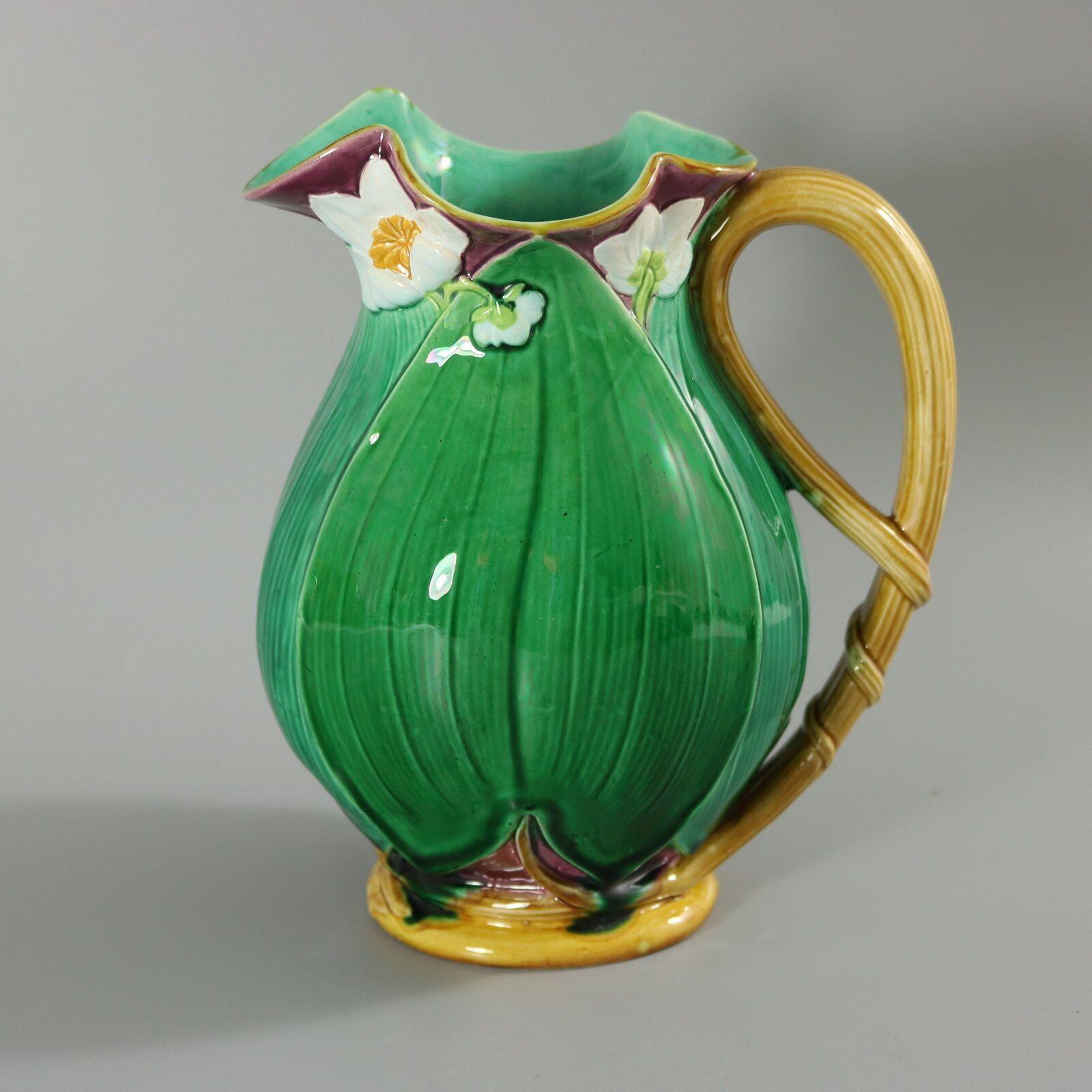 Minton Majolica jug/pitcher which features flowering lilies and lily pads. Colouration: green, white, dark pink, are predominant. The piece bears maker's marks for the Minton pottery. Bears a pattern number, '1228 12'. Marks include a factory