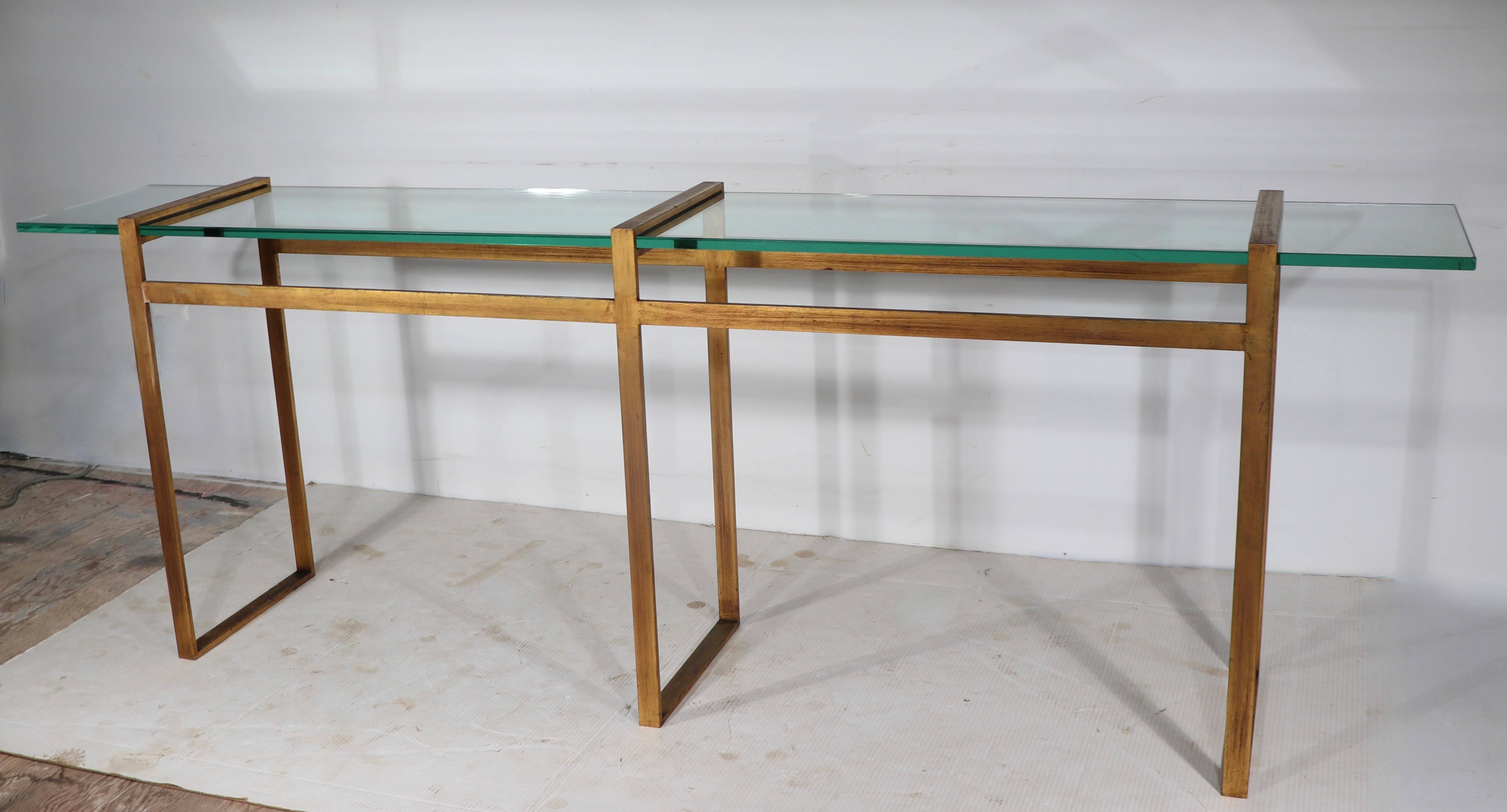 Voguish, sophisticated and chic architectural console table, having a squared metal frame in faux gilt finish, with a large, thick ( .75 in ) glass shelf. 
The console is in very good original condition showing only light cosmetic wear normal and