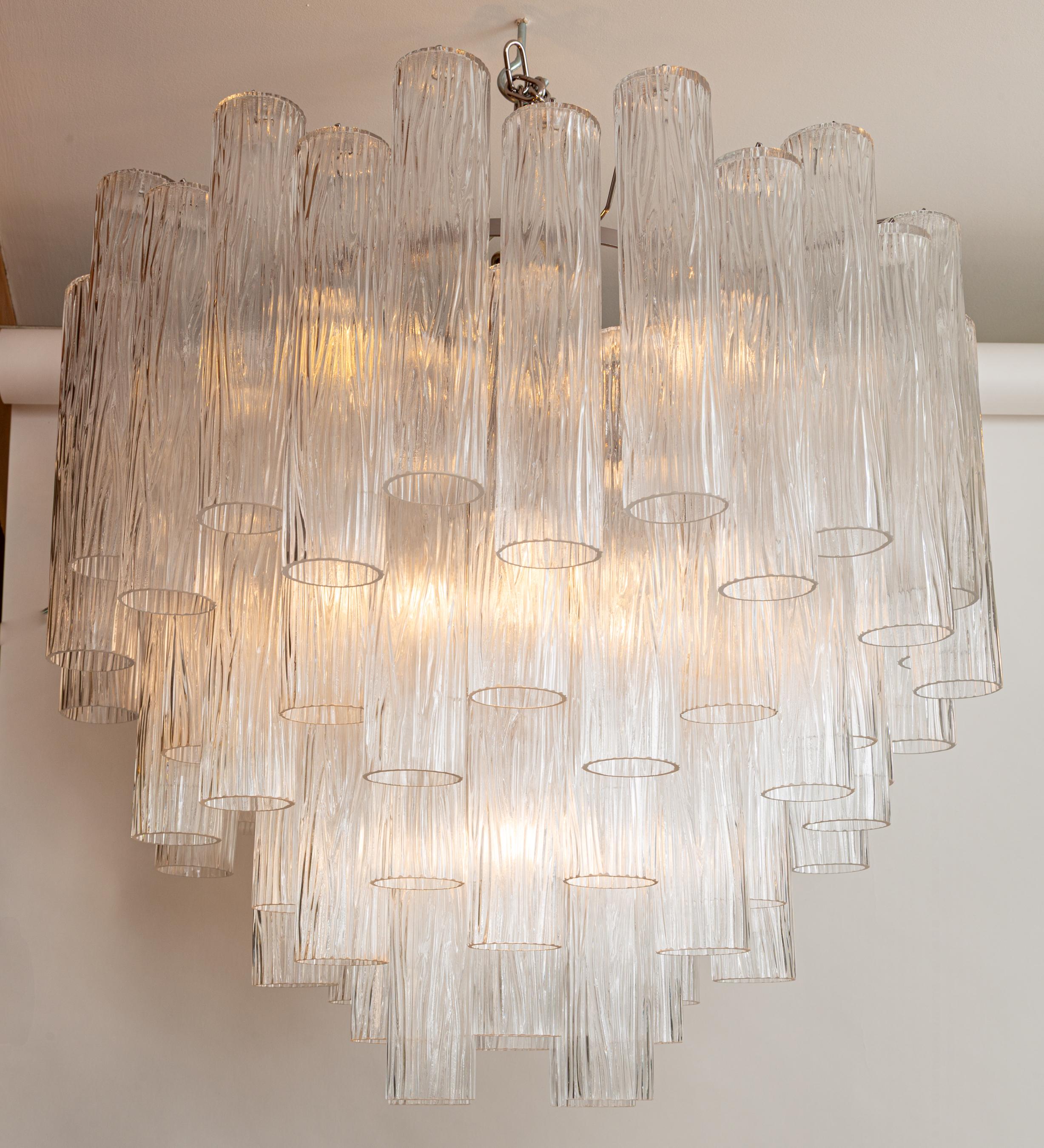 Un lampadario con struttura in nichel dalle linee eleganti, composto da quattro livelli di tubi cilindrici in vetro soffiato trasparente sfalsati, con un design dalla texture naturalistica. Composto da oltre 80 pezzi di vetro.
Venduto come