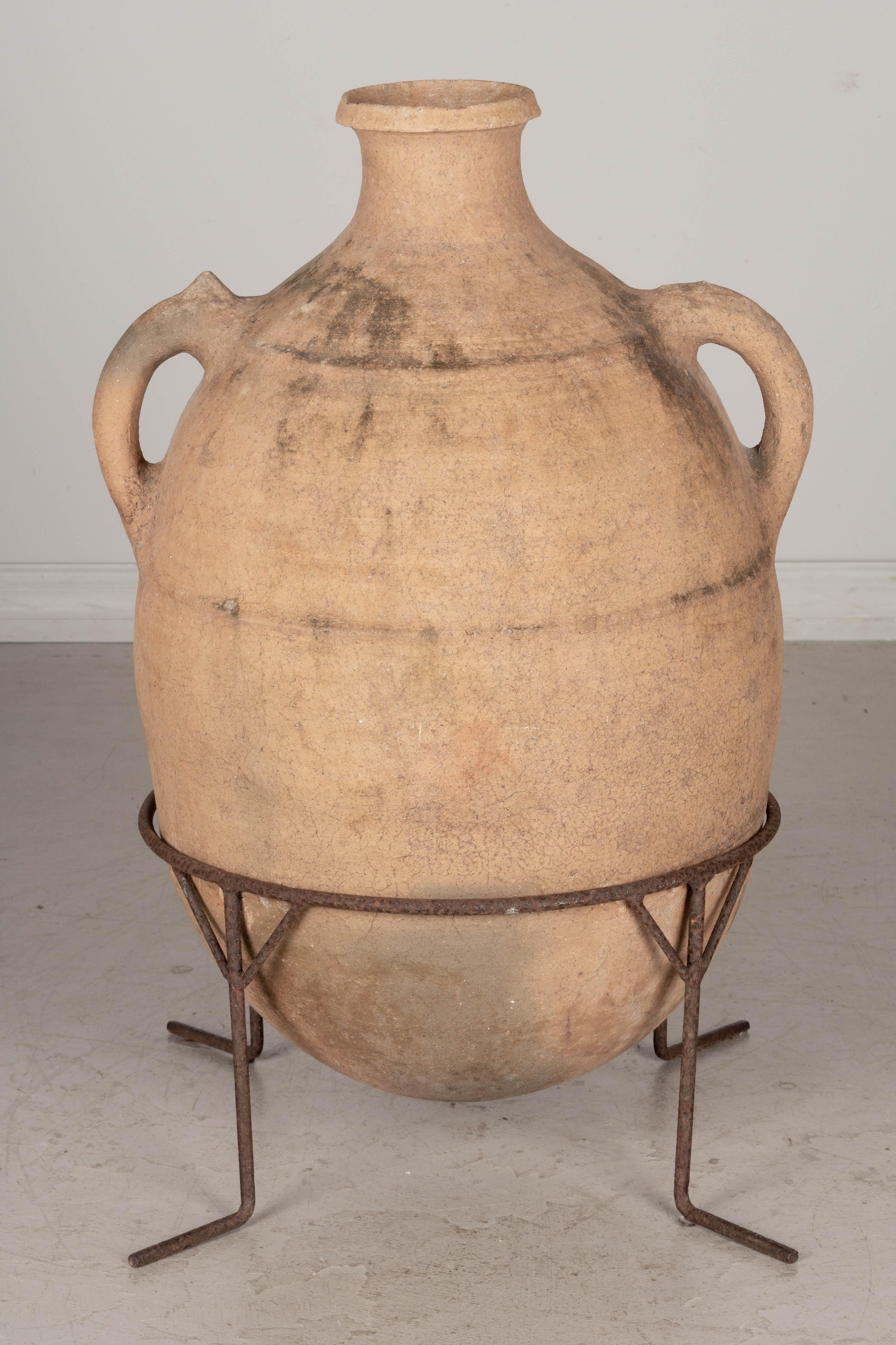A large Moroccan terracotta pottery water jar resting in an iron stand. Oval form with rounded bottom and two small handles. Iron has rusty patina. Minor chips. Beautiful clay color. 
Overall dimensions including stand: 28