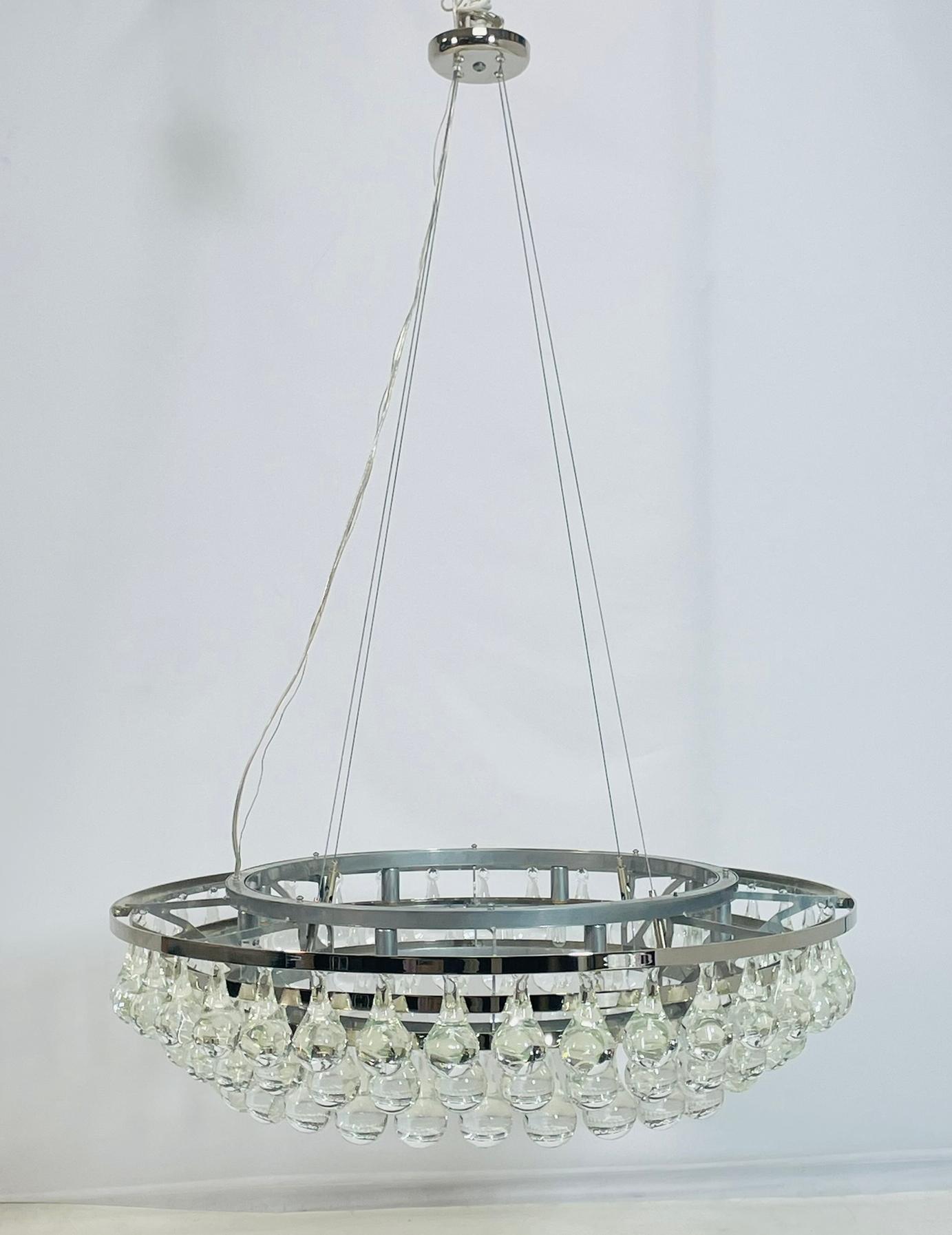 Esta elegante lámpara de araña presenta un marco circular de níquel pulido adornado con colgantes en forma de lágrima de cristal de Murano soplado a mano. Las gotas de cristal transparente están suspendidas en varias capas, creando un lujoso efecto