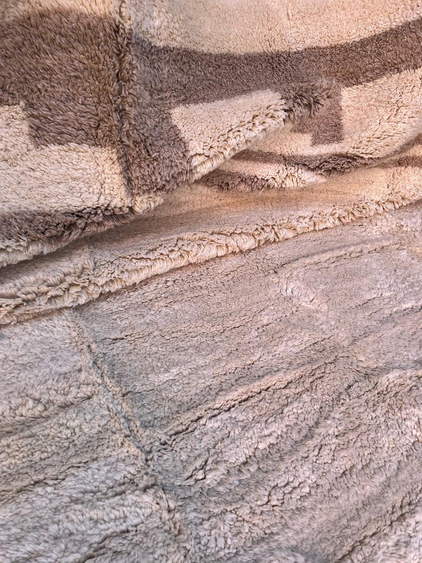 Ce grand tapis marocain neutre tissé à la main a été fabriqué par des artisans qualifiés dans les montagnes de l'Atlas. Il est composé de laine M'Rirt crème pelucheuse de haute qualité, avec une bordure unique dans une couleur taupe neutre. Ce tapis