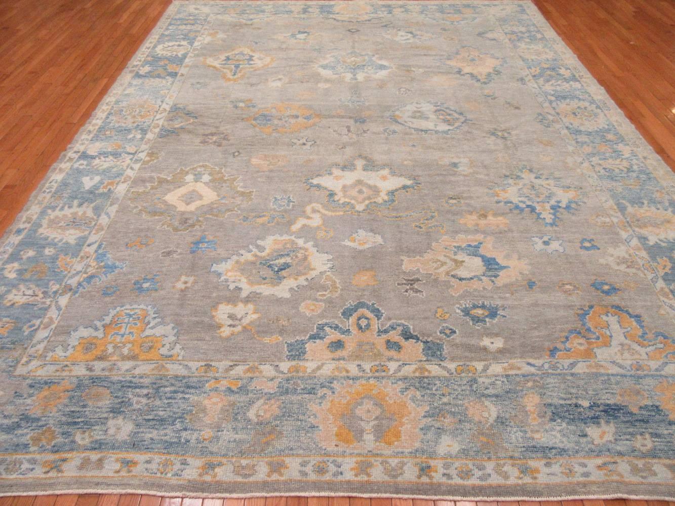 This is a large new hand-knotted wool rug from Turkey woven in the infamous Oushak design. It is a well constructed rug with primary colors of ivory, gray, blue and orange red. Its transitional design makes it an easy rug to work with for any room