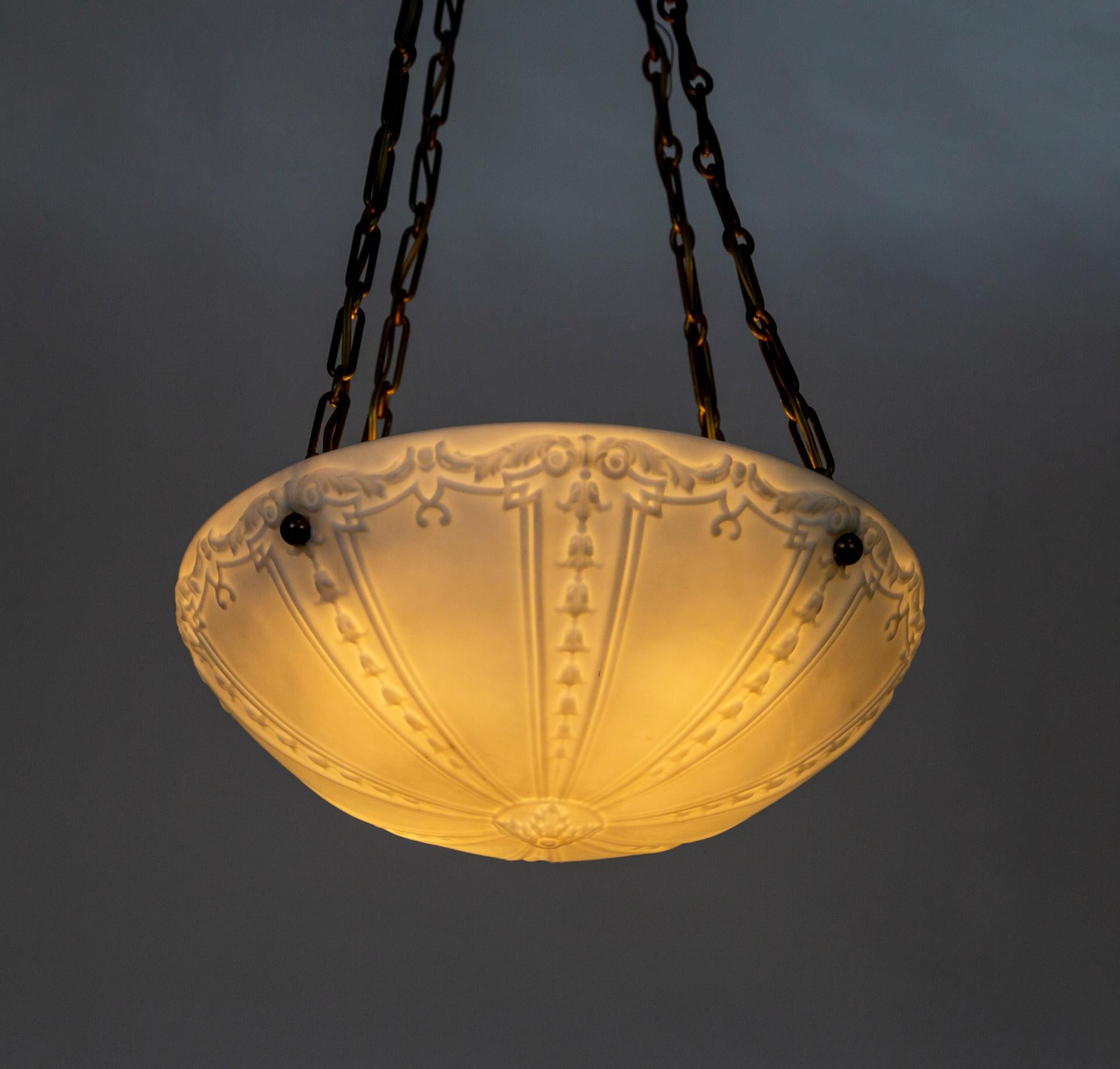 A pair of large, hanging, up-lights with white, glass, bowl shades from the early 20th century. The imprinted glass is detailed with acanthus leaf garlands and radial bell flowers. They hang from four long, decorative chains and etched brass