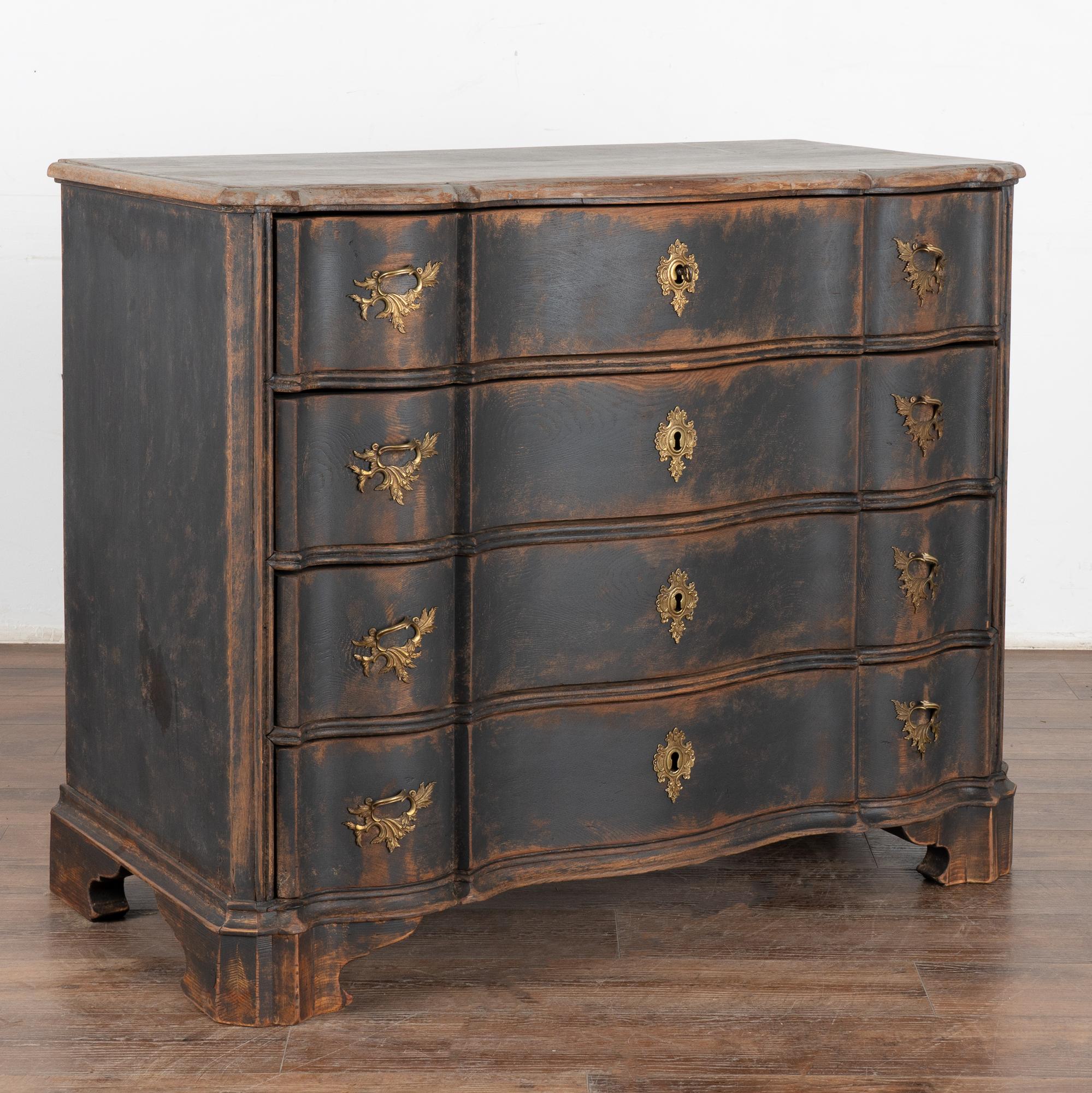 This antique rococo large oak chest of drawers features a serpentine front, brass hardware pulls and is raised on carved feet. 
The newer, professionally applied and layered black painted finish is gently scraped and distressed, revealing the