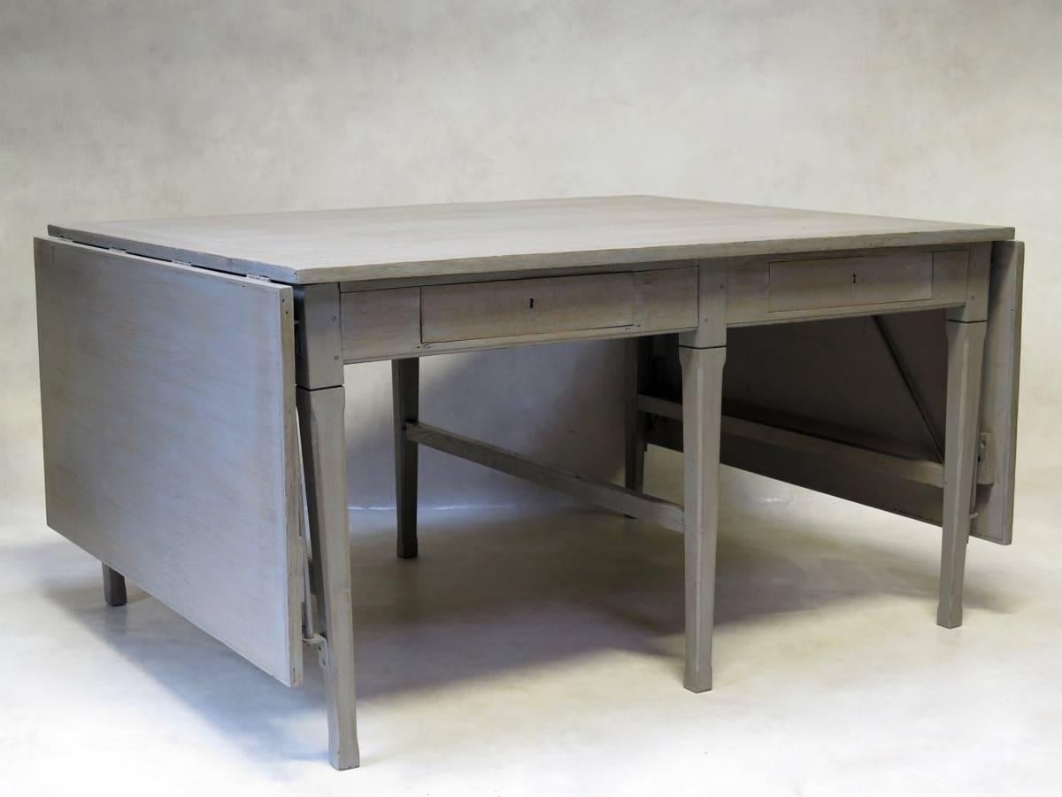 Heavy, solid oak work table, from the French postal service, of efficient, pared-down design. The table has a light-grey, cerused-like finish, and folding extensions on either end. The table has two drawers on one side, and one on the opposite side.