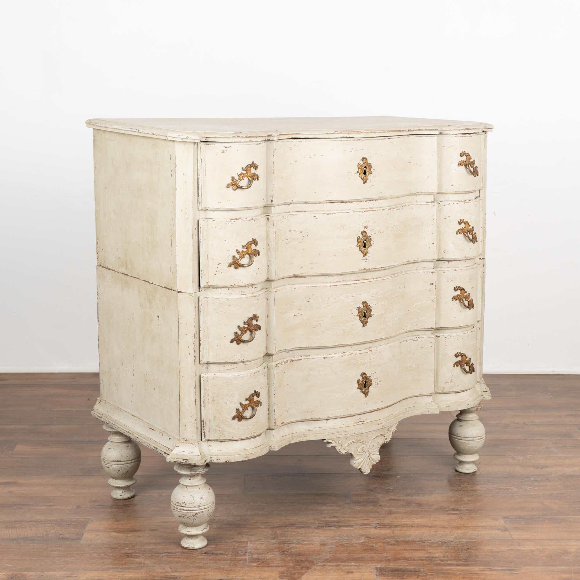 This antique rococo large oak chest of drawers features a serpentine front, brass hardware pulls and is raised on large turned feet.
The newer, professionally applied and layered white painted finish has soft gray undertones and is gently scraped